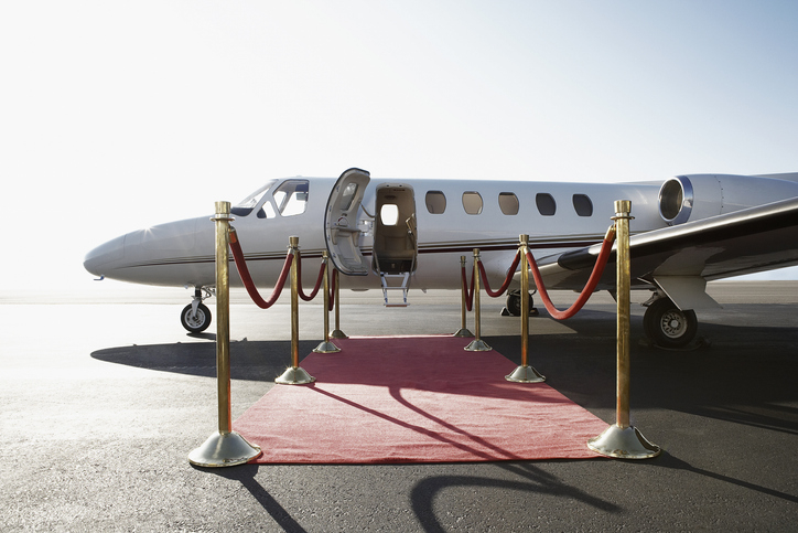 A private jet with its door open is parked on an airport runway, accessed by a red carpet flanked by gold stanchions and ropes