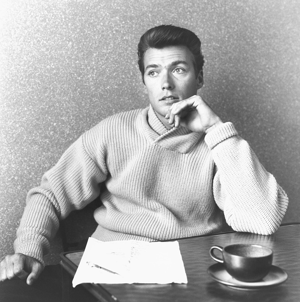 Clint Eastwood sits at a table with one hand under his chin, wearing a knit sweater, with a sheet of paper and a cup of coffee in front of him