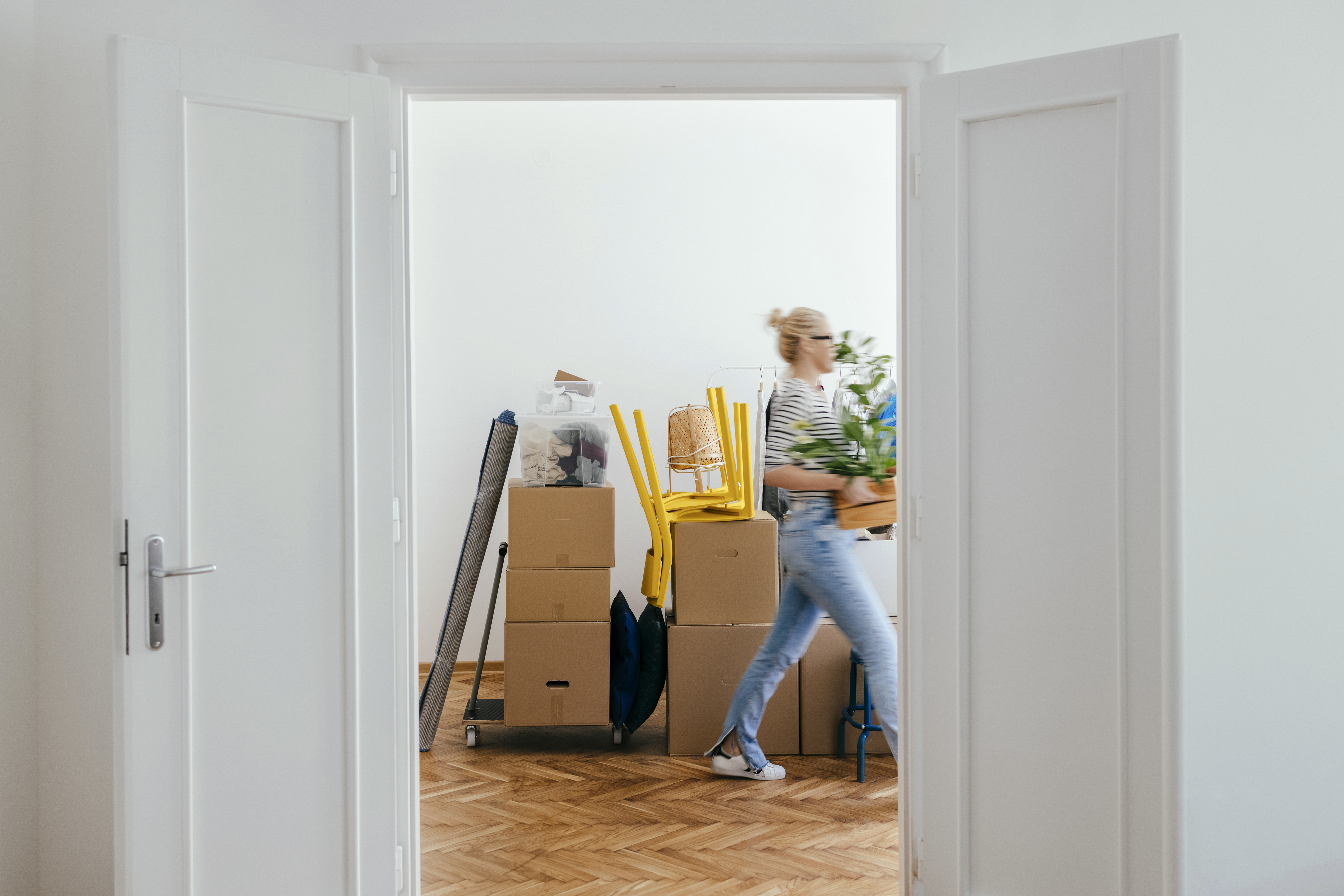 Person carrying a plant walks through a doorway in a room filled with moving boxes and furniture