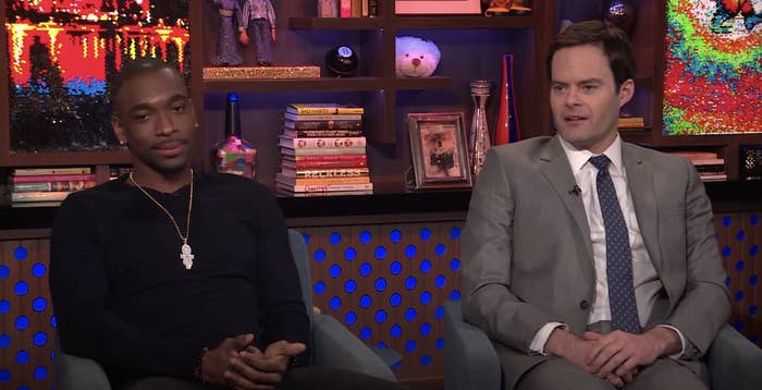 Jay Pharoah in a black shirt and Bill Hader in a gray suit are seated and talking on a talk show set with a colorful background
