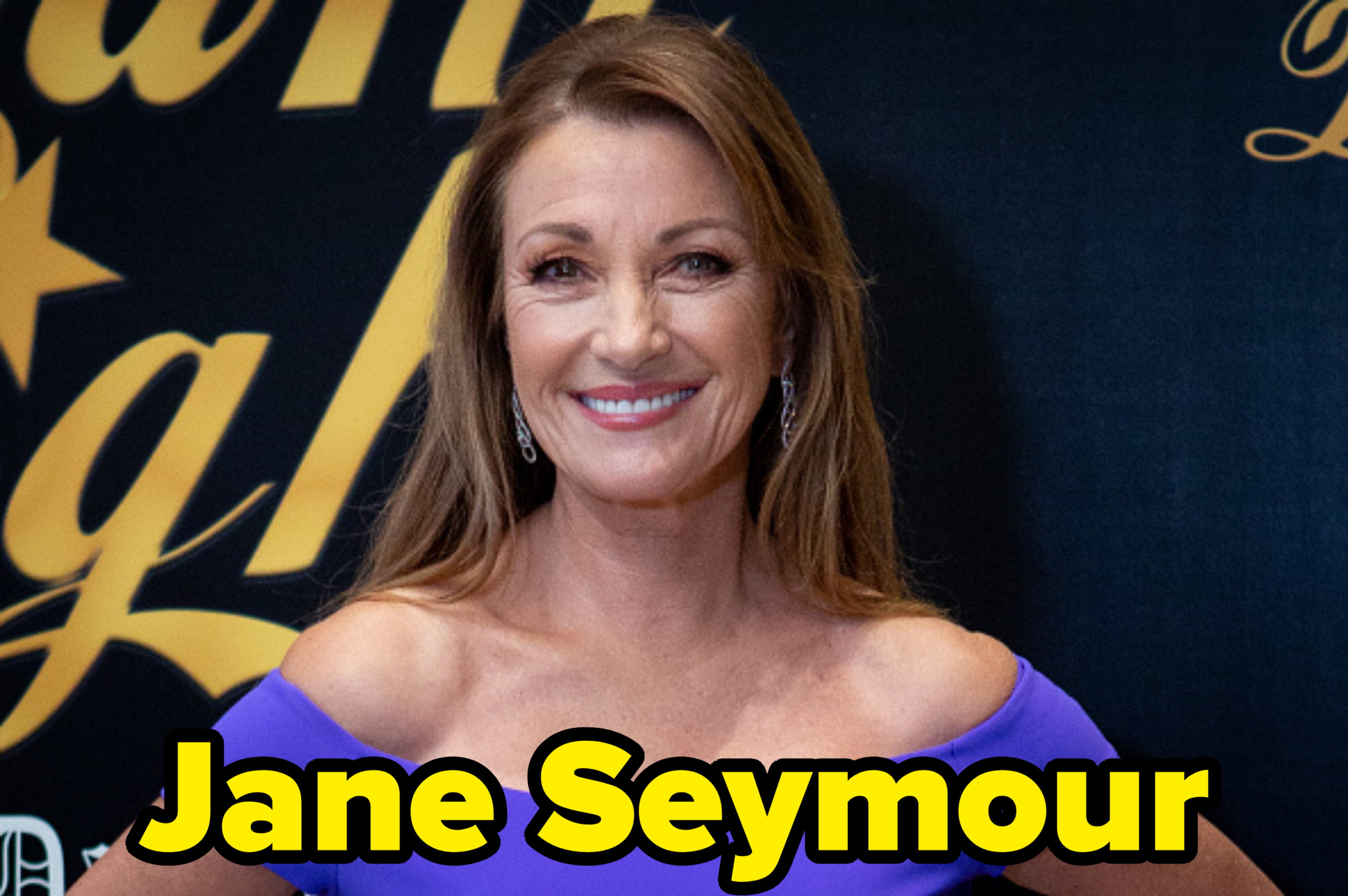 Jane Seymour smiles wearing an off-the-shoulder dress with dangling earrings, in front of a backdrop with cursive text