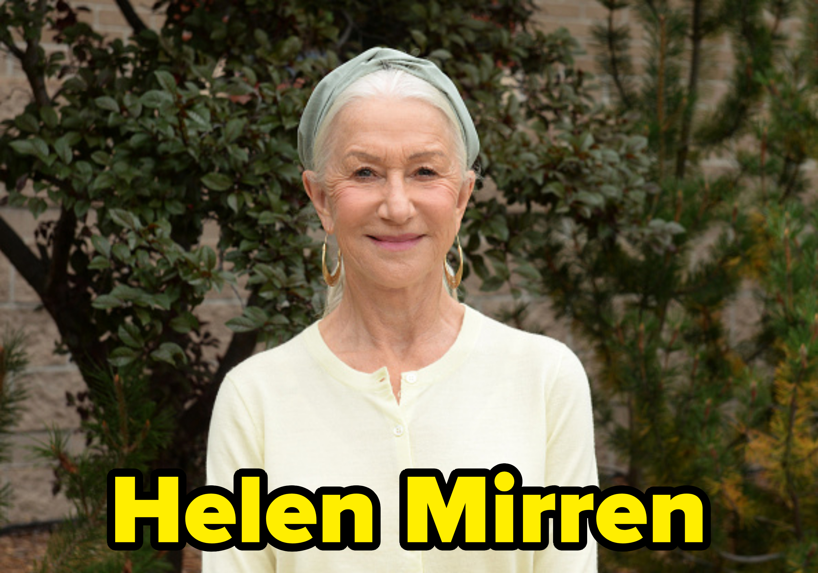 Helen Mirren smiling outdoors, wearing a light cardigan and headwrap with hoop earrings, standing in front of greenery