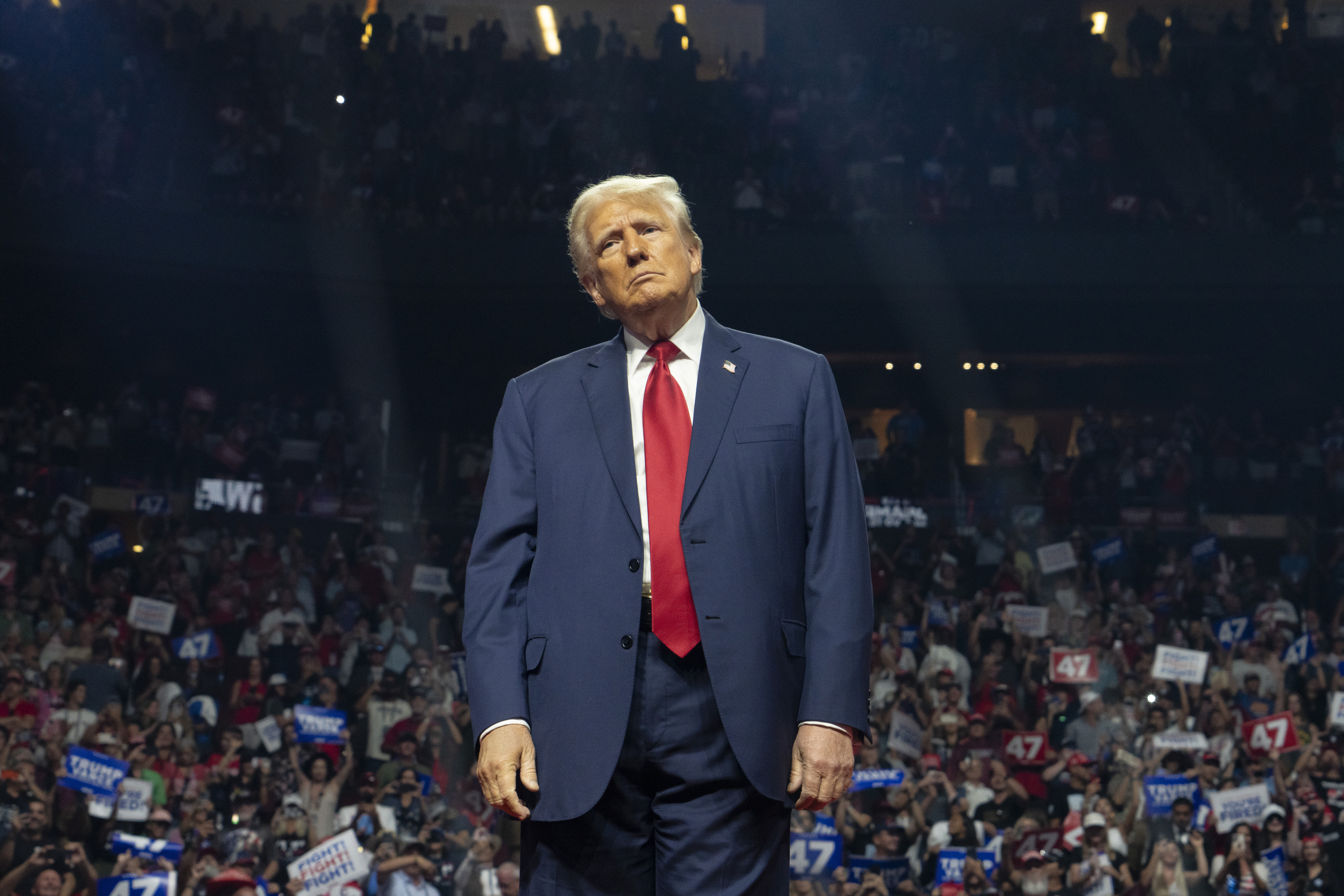 Donald Trump stands on a stage at a rally, wearing a suit with a red tie. Audience members hold up signs and banners in the background