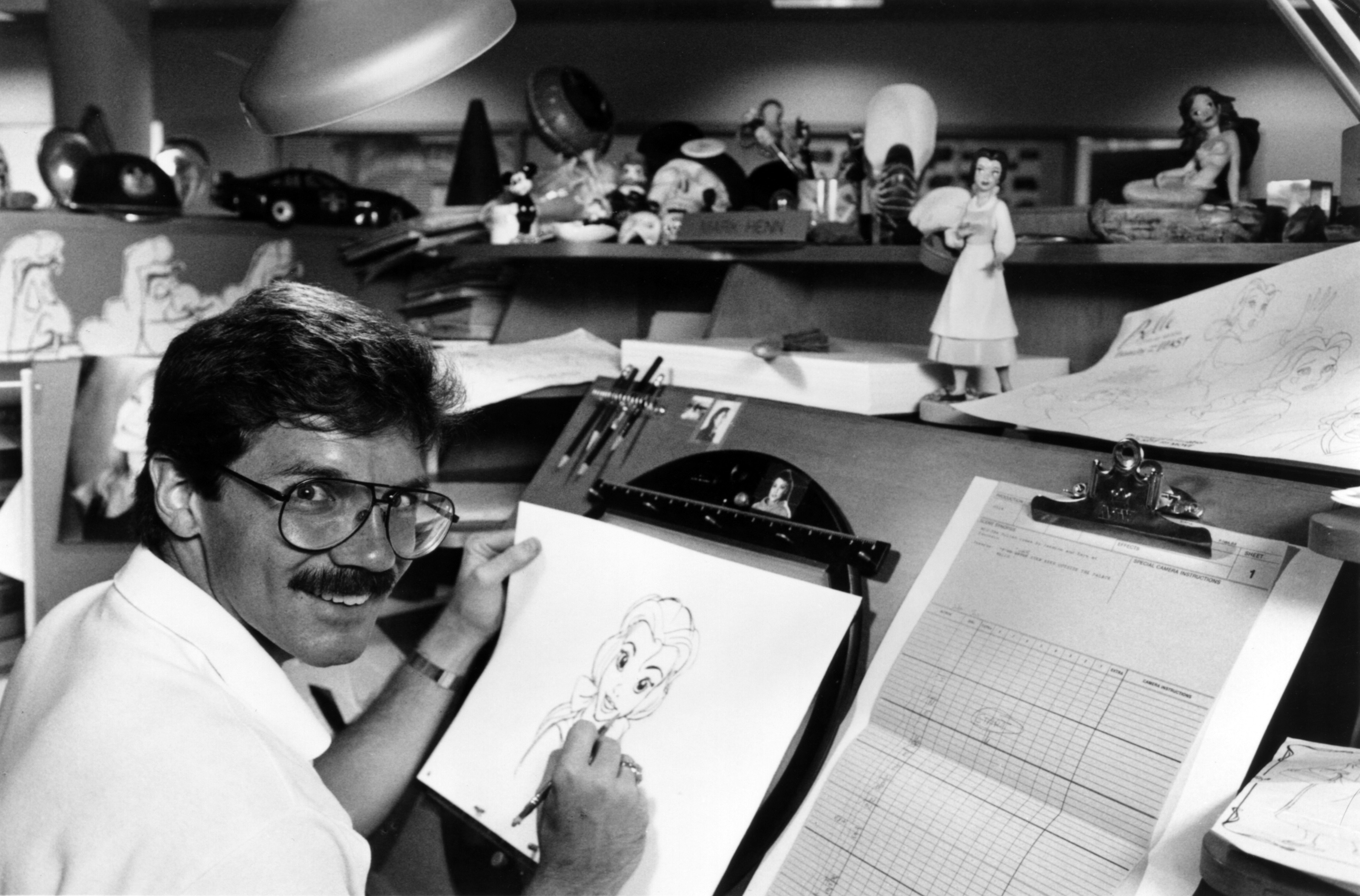 A man is drawing a cartoon character at a desk filled with animation tools, sketches, and character figurines in an animation studio
