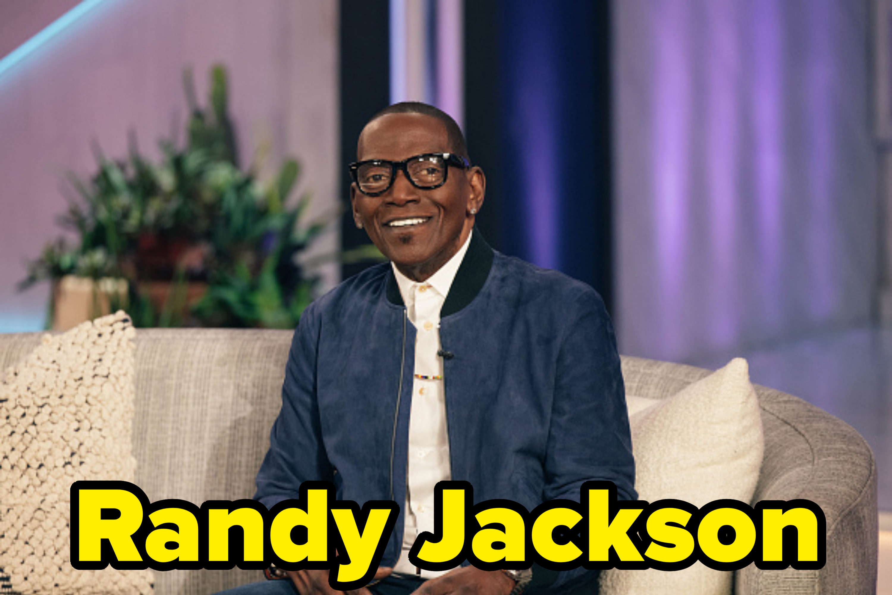 A person is sitting on a couch wearing glasses, a casual jacket, and smiling