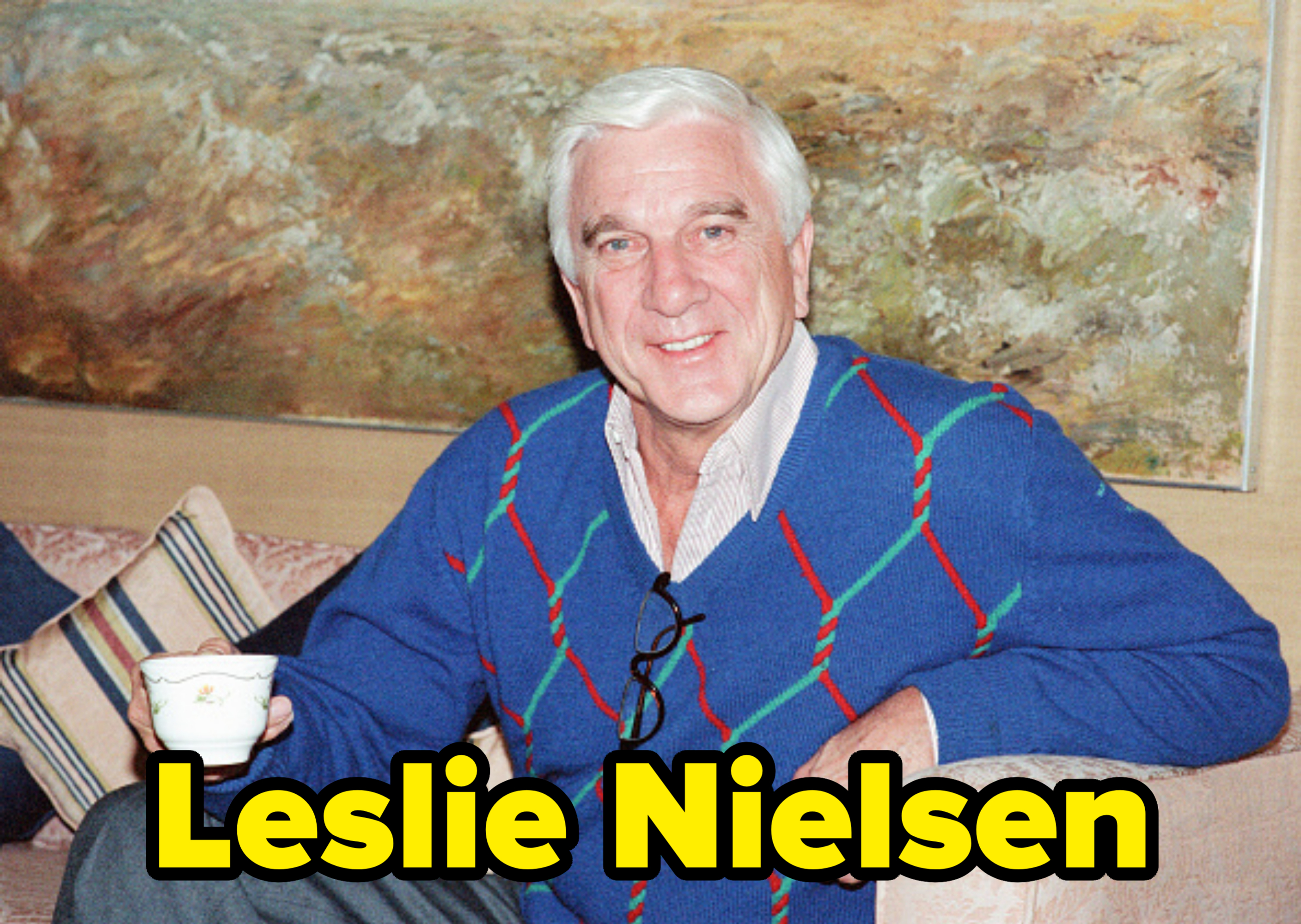 An elderly man in a blue patterned sweater, holding a teacup, smiles while seated on a couch