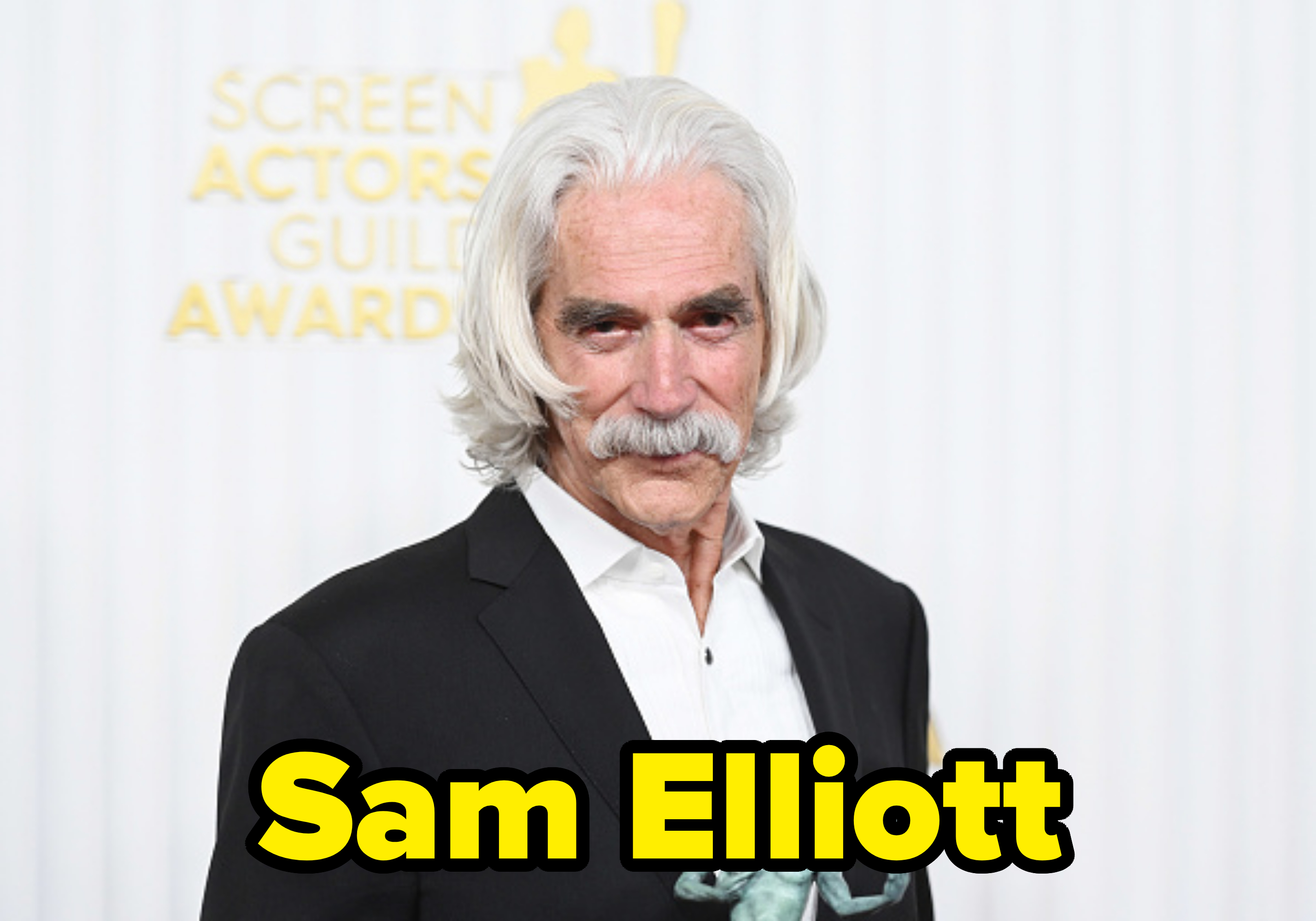 An older man with long white hair and a mustache, dressed in a black suit and white shirt, holds a statuette at the Screen Actors Guild Awards