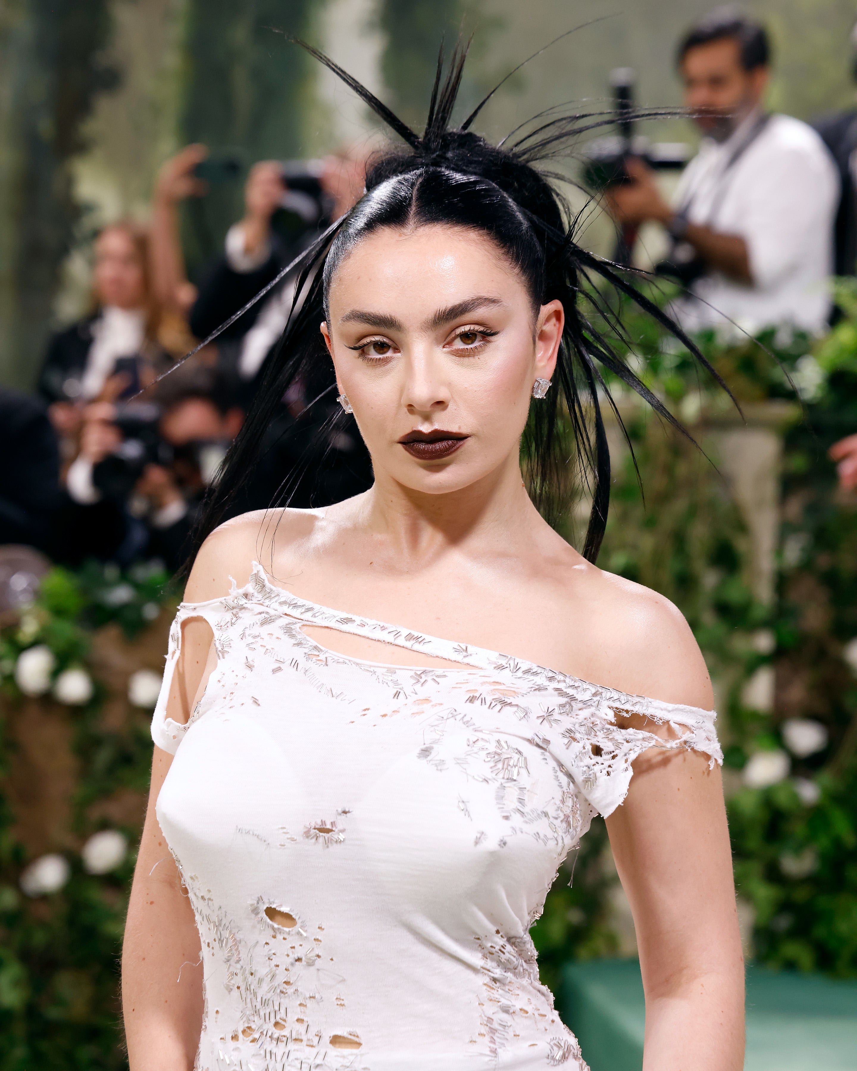 Charli XCX at a fashion event in an asymmetrical, distressed dress with cutouts, and her hair styled in an avant-garde, spiky updo