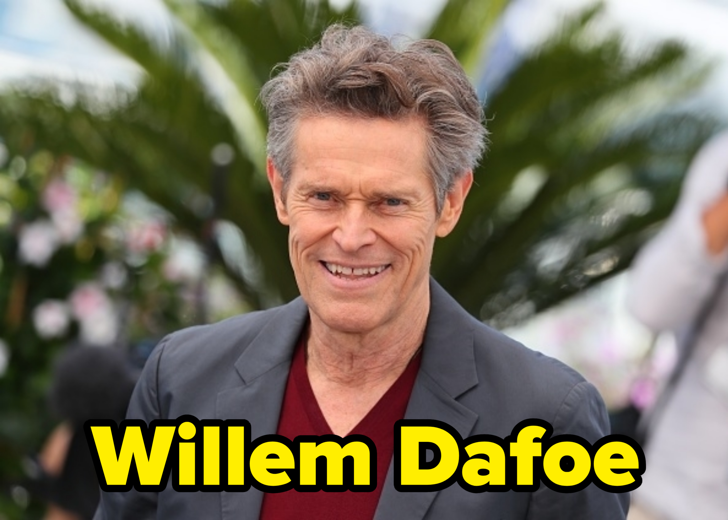 Willem Dafoe smiles while wearing a blazer over a V-neck top, standing in front of a blurred foliage background