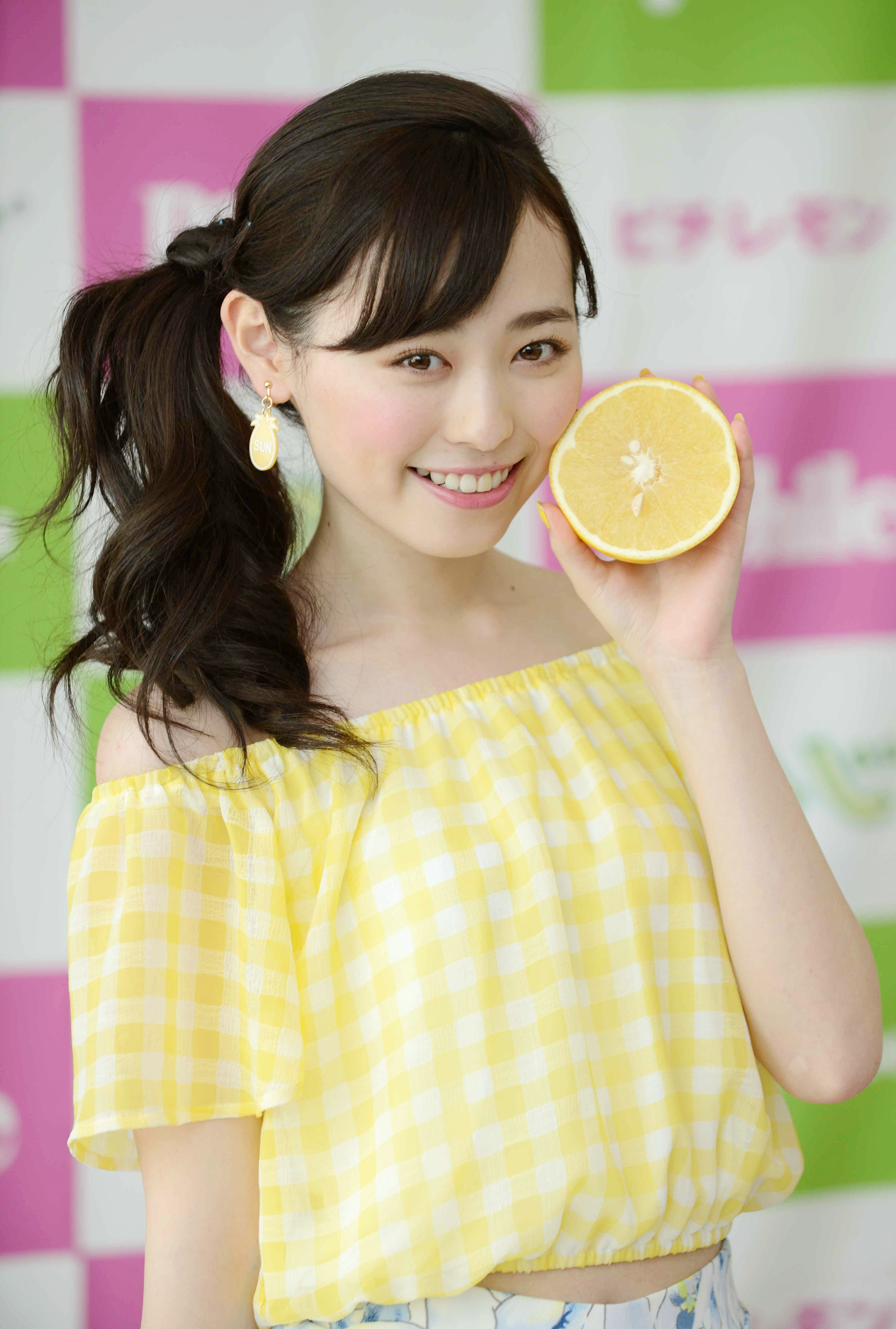 女性が黄色のチェック柄のオフショルダーブラウスを着て片手に半分に切ったオレンジを持ち、笑顔を見せている。名前は不明。