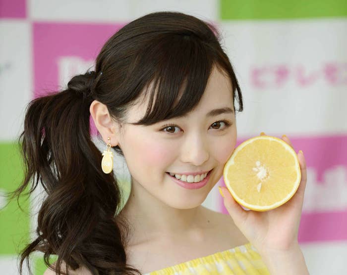 女性が黄色のチェック柄のオフショルダーブラウスを着て片手に半分に切ったオレンジを持ち、笑顔を見せている。名前は不明。