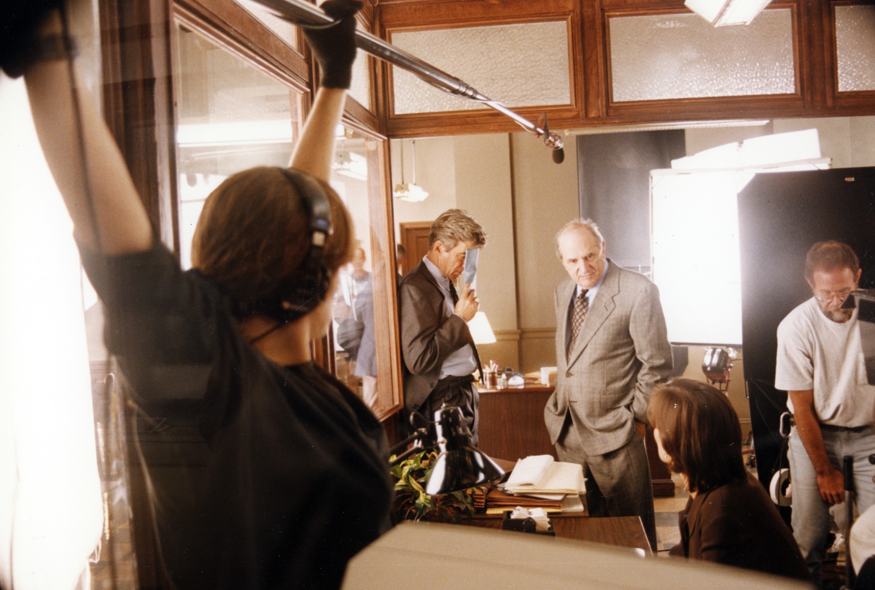 Behind-the-scenes on a TV/film set: crew with equipment and actors in suits surrounded by lighting and cameras, engaged in discussion