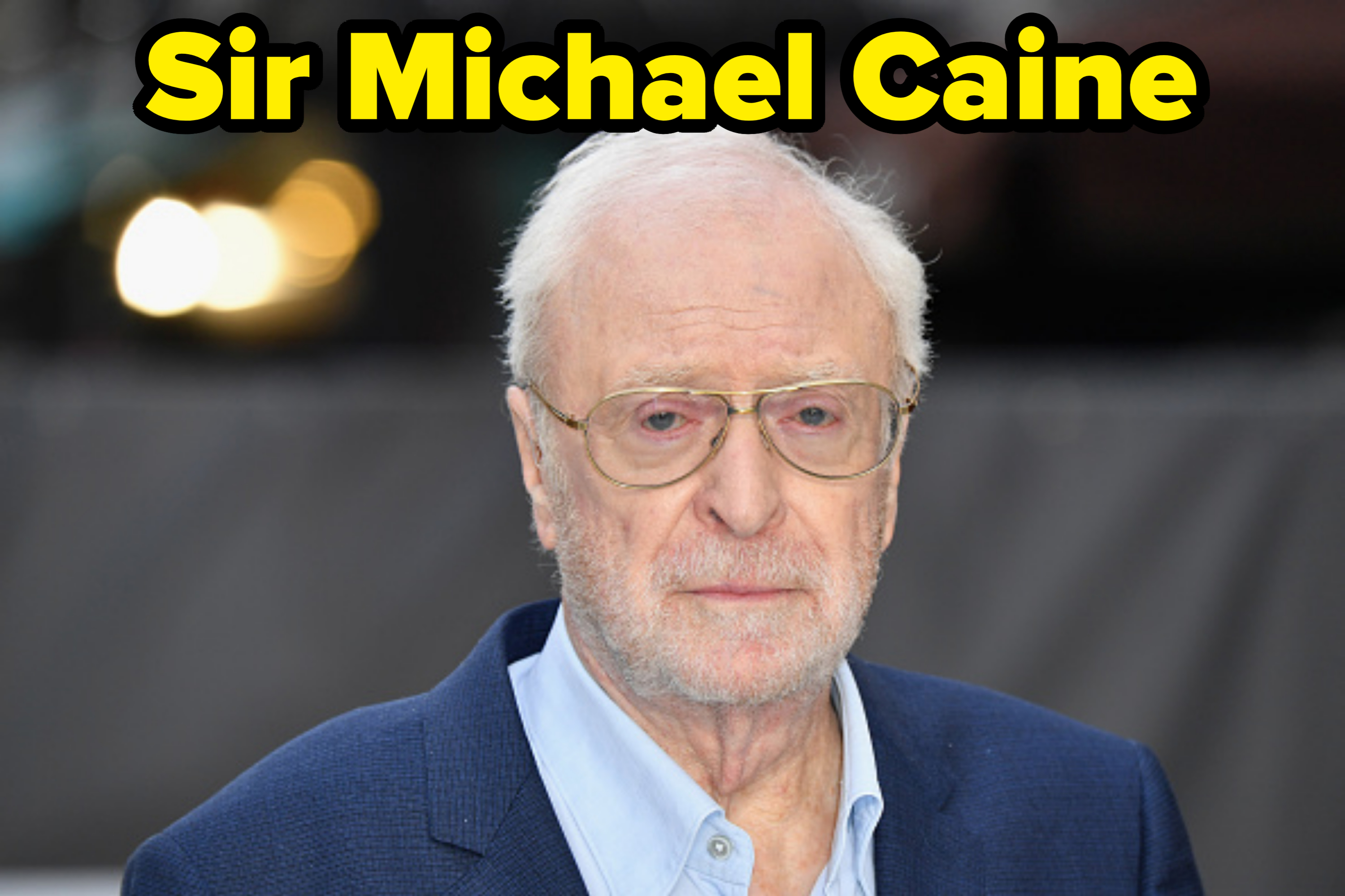 Michael Caine wearing a dark blue blazer over a light blue shirt, with glasses, looking at the camera