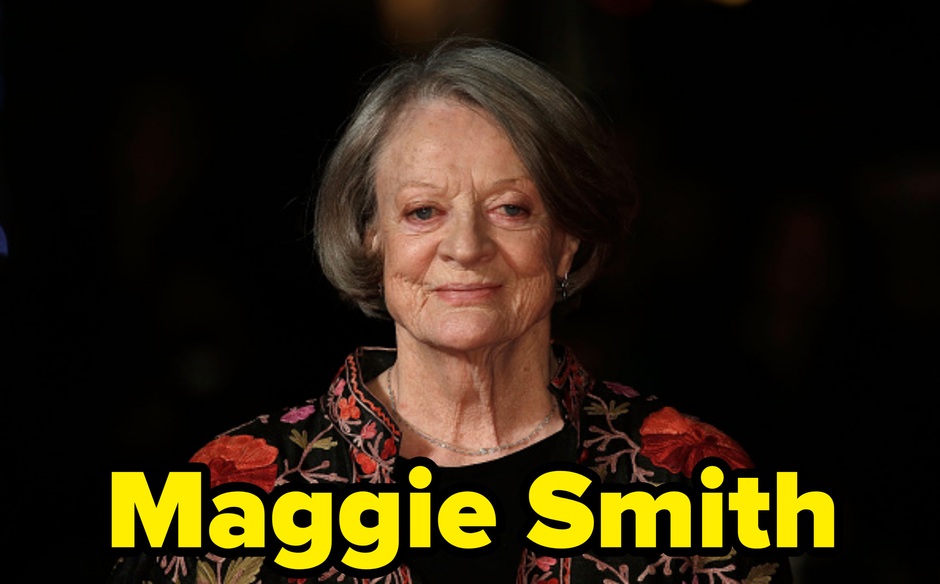 Maggie Smith stands on the red carpet, wearing a floral-patterned outfit, looking directly at the camera with a neutral expression