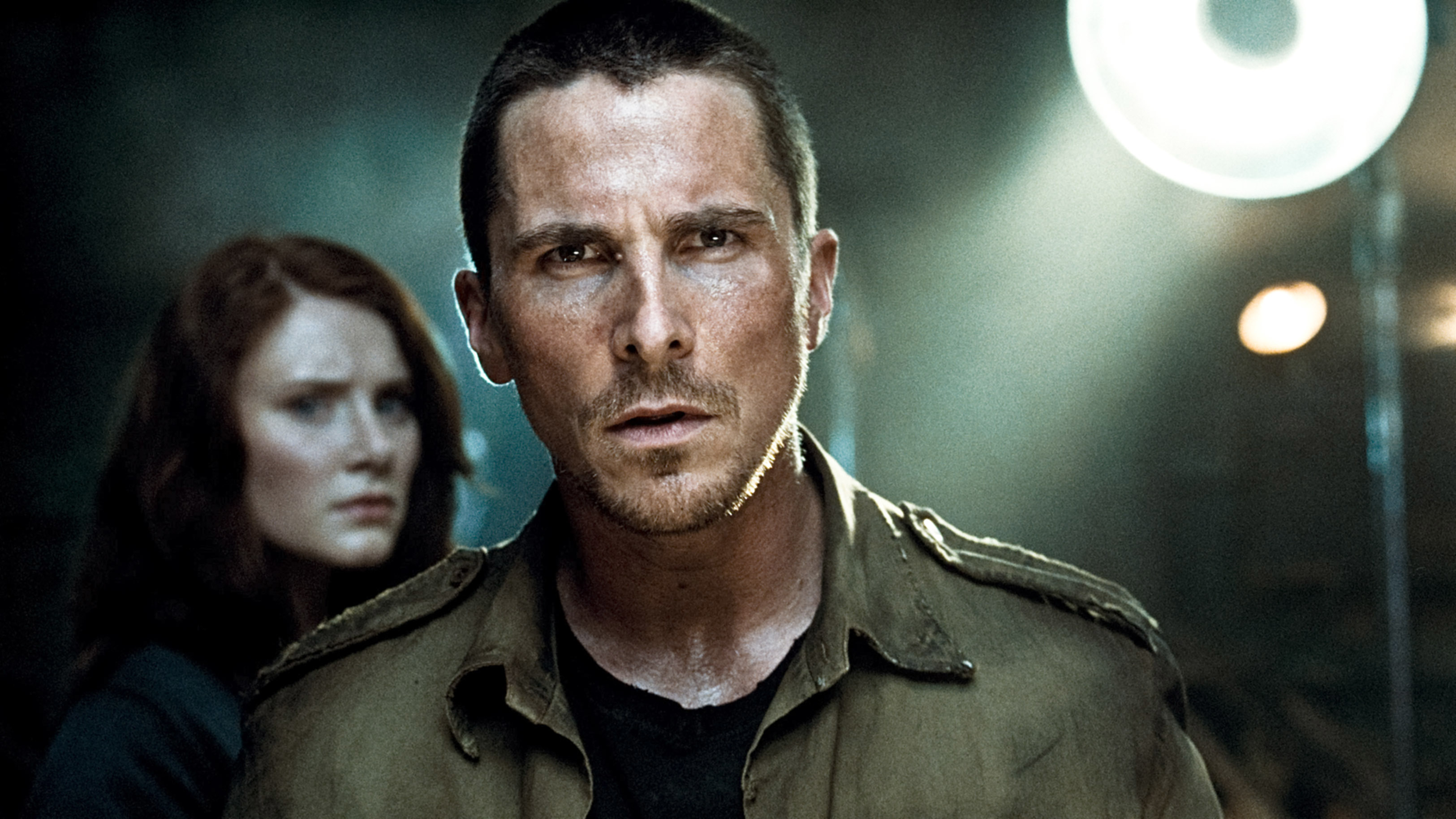 Christian Bale wearing a military-style jacket, looking intense, with Bryce Dallas Howard blurred in the background. Both appear in a dramatic scene