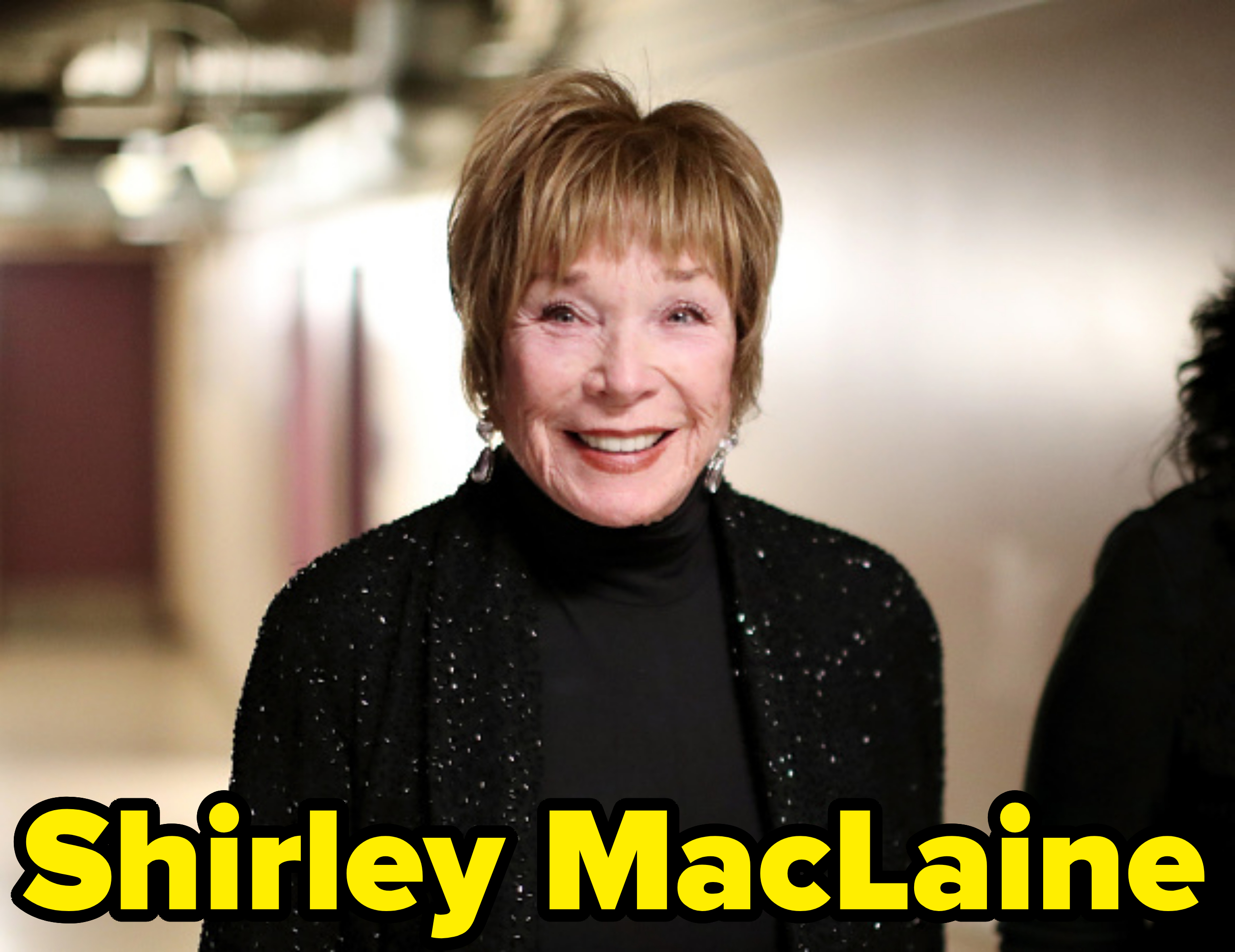 Shirley MacLaine smiles while wearing a black, sparkly blazer over a dark turtleneck. The background is slightly blurred