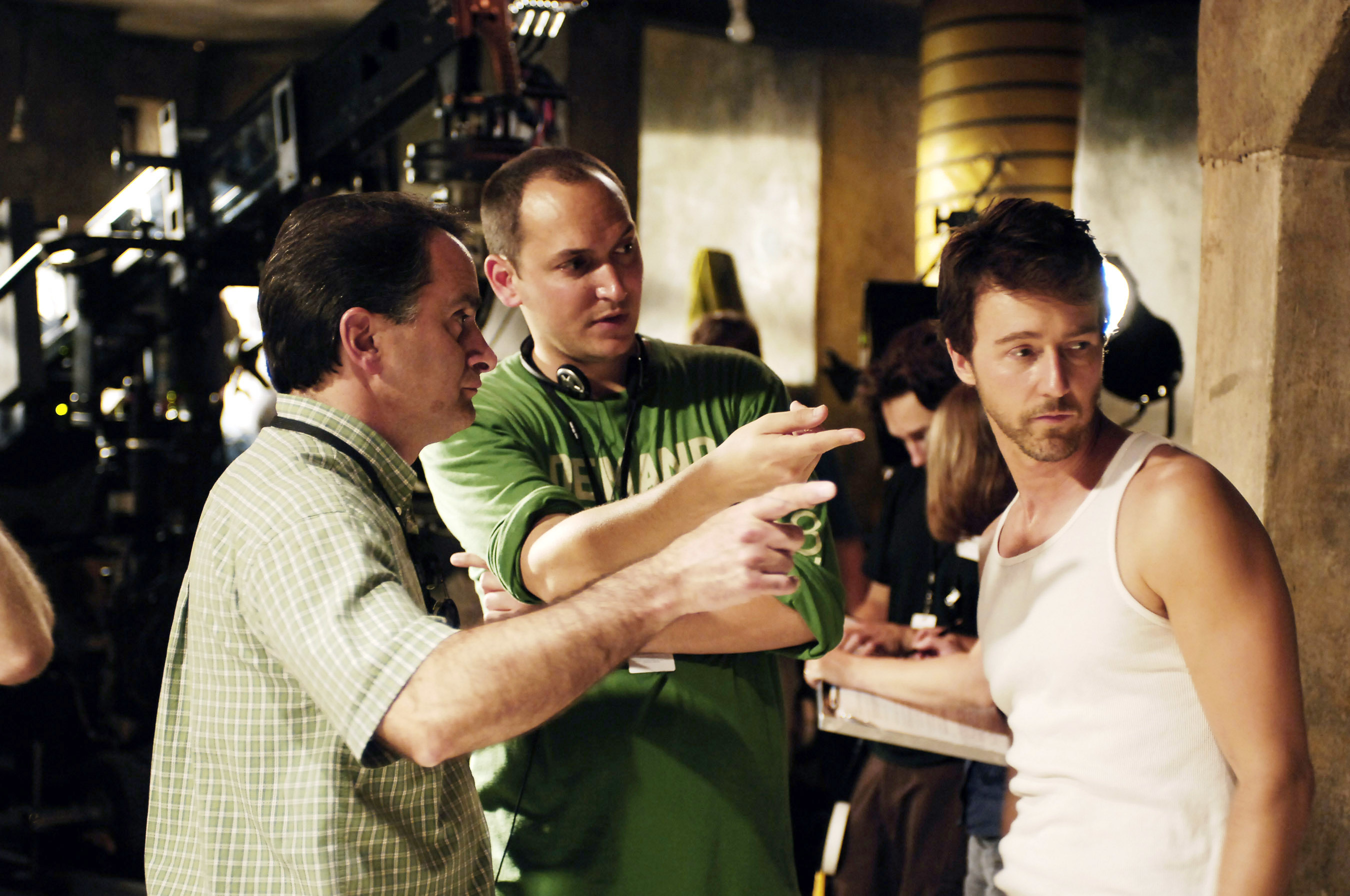 Three men, including director Louis Leterrier and Edward Norton, in a casual conversation on a film set. Edward is wearing a white tank top; the other two men appear to be discussing the scene