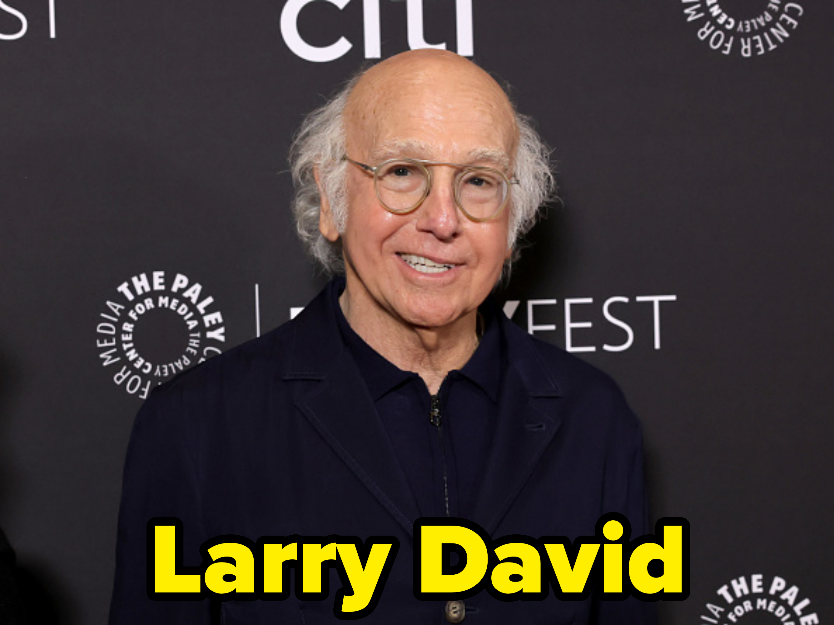 Larry David at a PaleyFest event, wearing a dark suit with glasses, and smiling at the camera