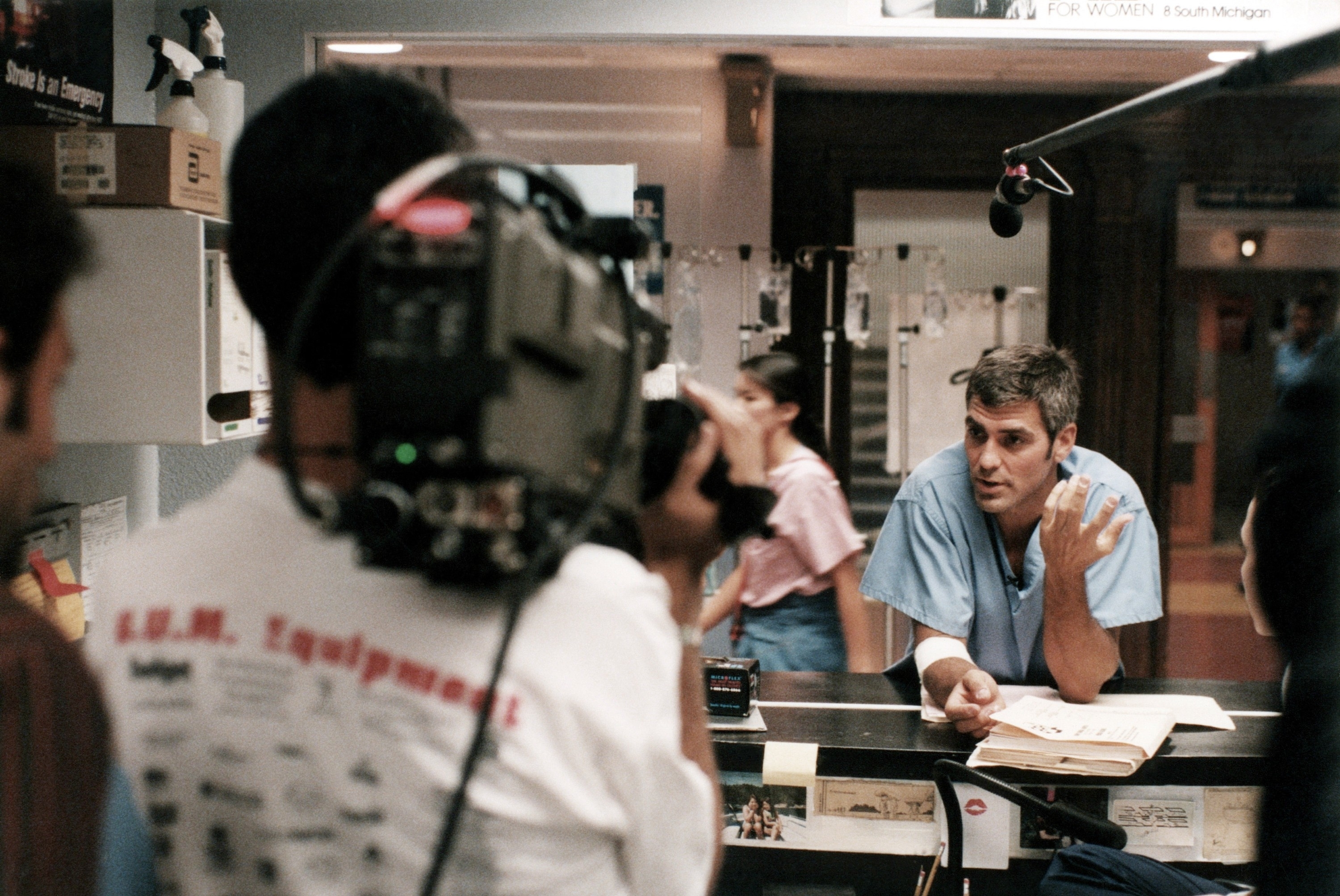 George Clooney is behind the scenes on a TV show set, wearing scrubs, engaging with the crew as a camera records him