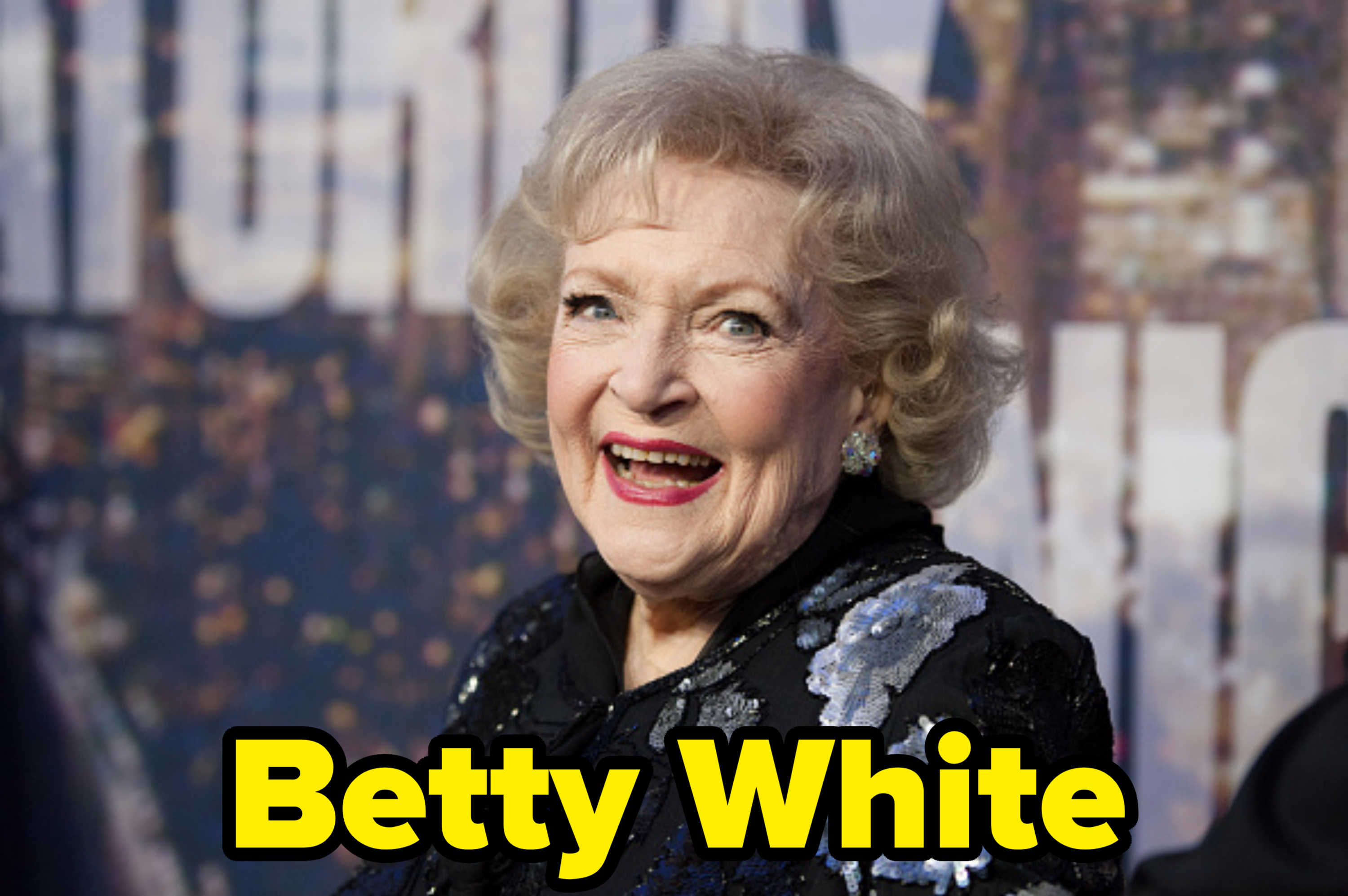 Betty White smiles broadly at an event, wearing a black outfit adorned with silver floral embellishments