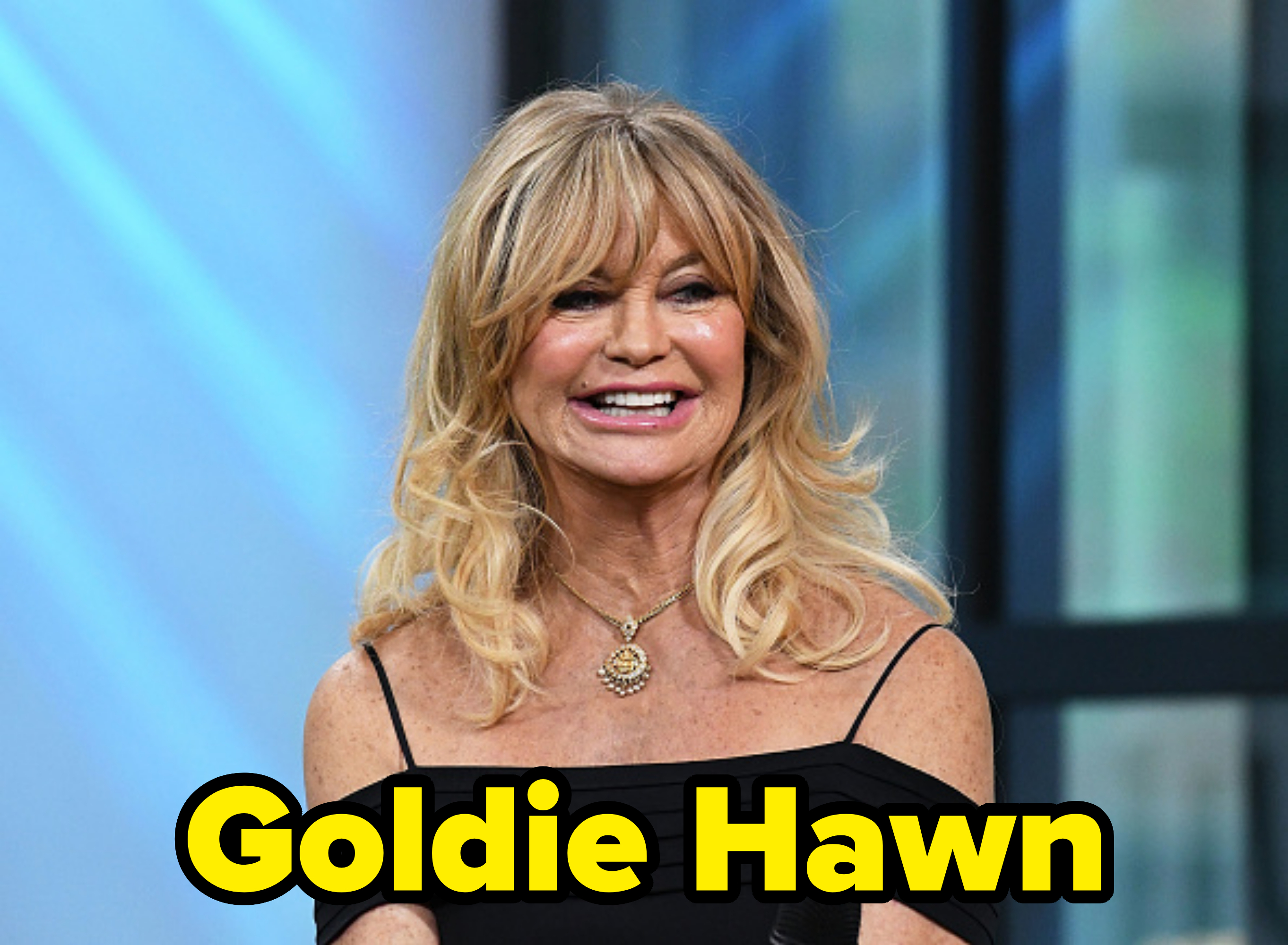 Goldie Hawn smiling, wearing an off-the-shoulder black dress and a gold necklace, holding a microphone