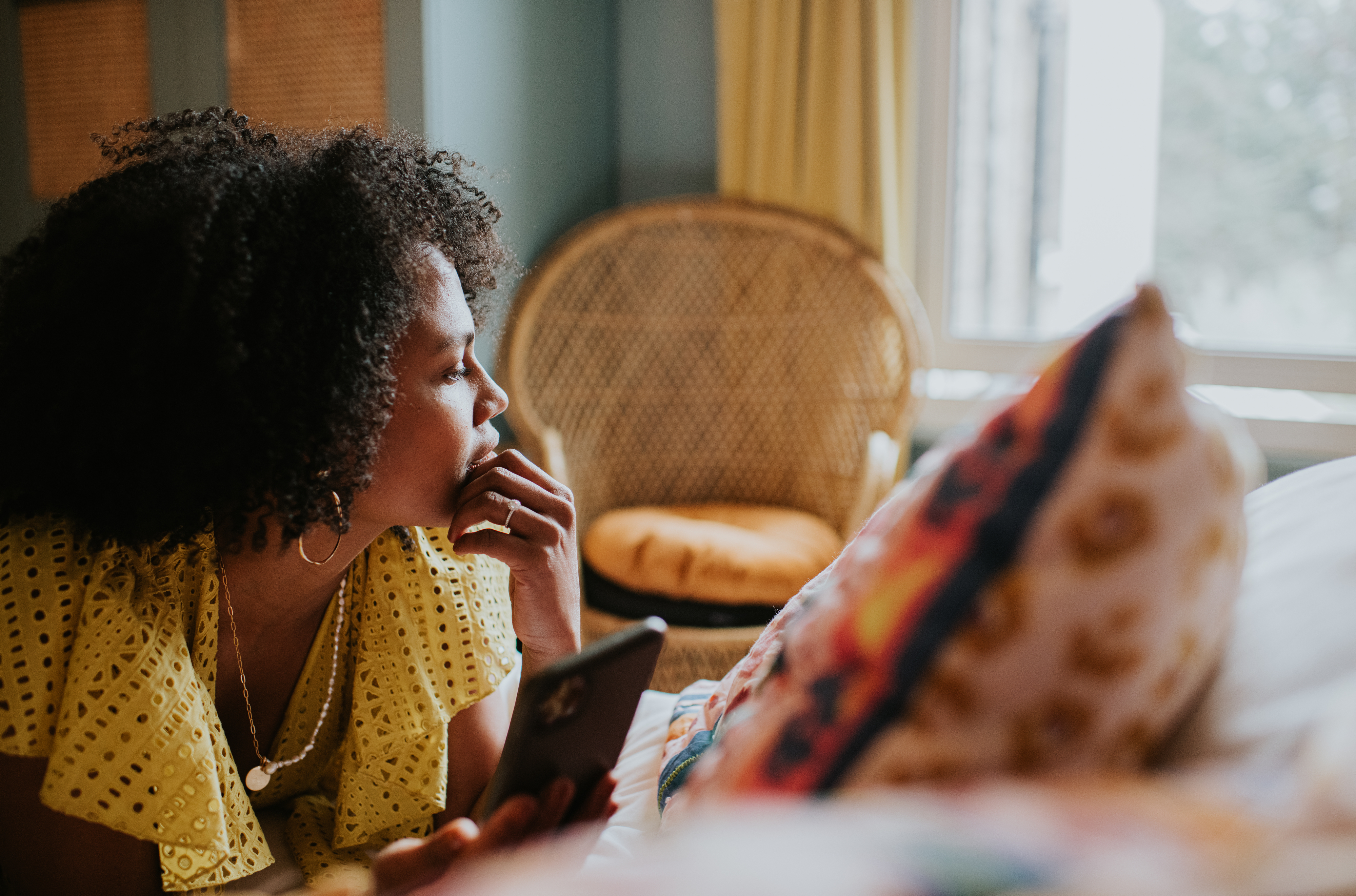 Woman in a yellow blouse looking thoughtful while holding a phone in a cozy, homey space