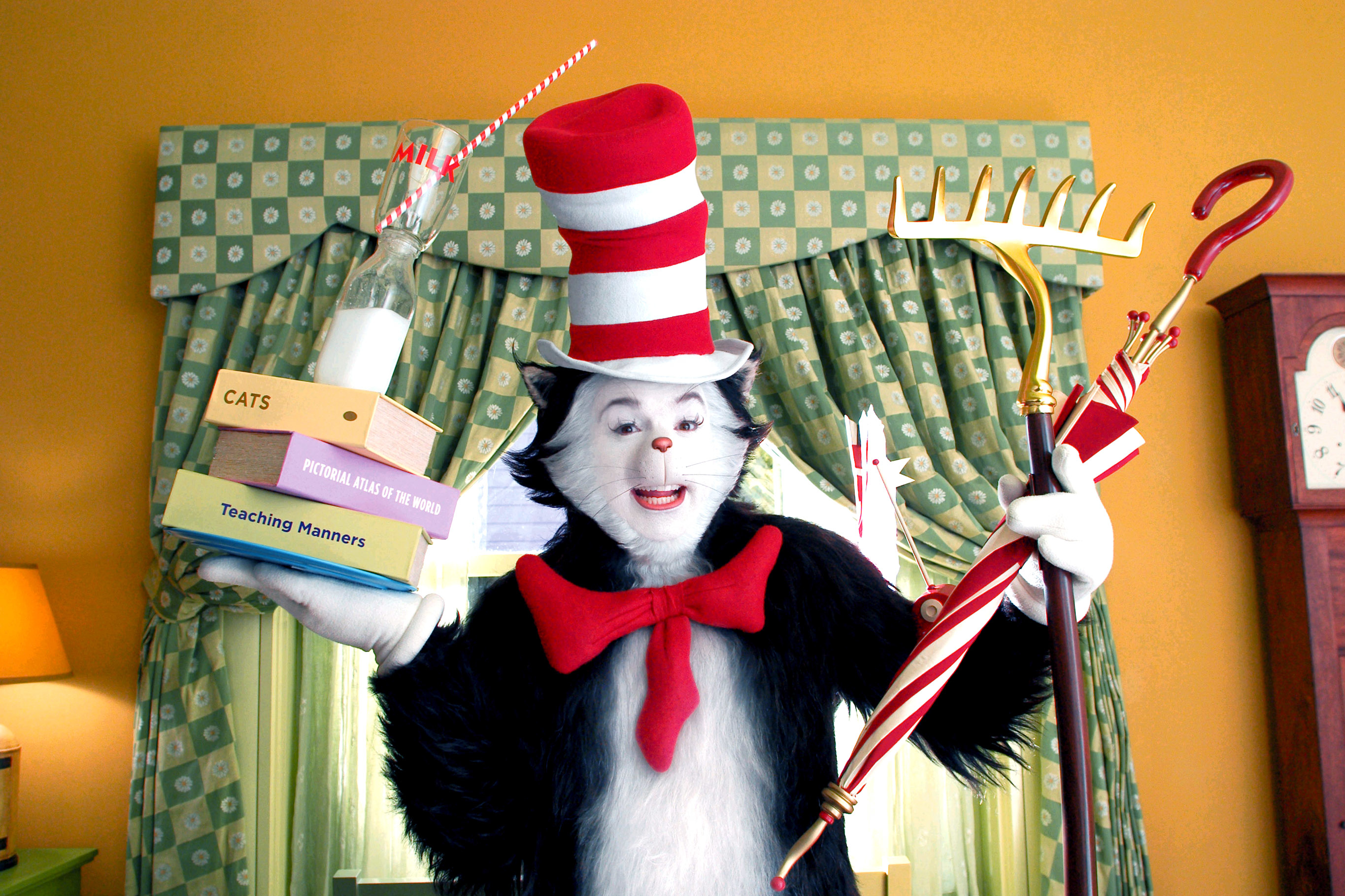 Mike Myers as The Cat in the Hat character holding a stack of books and a rake, in front of a polka-dotted curtain