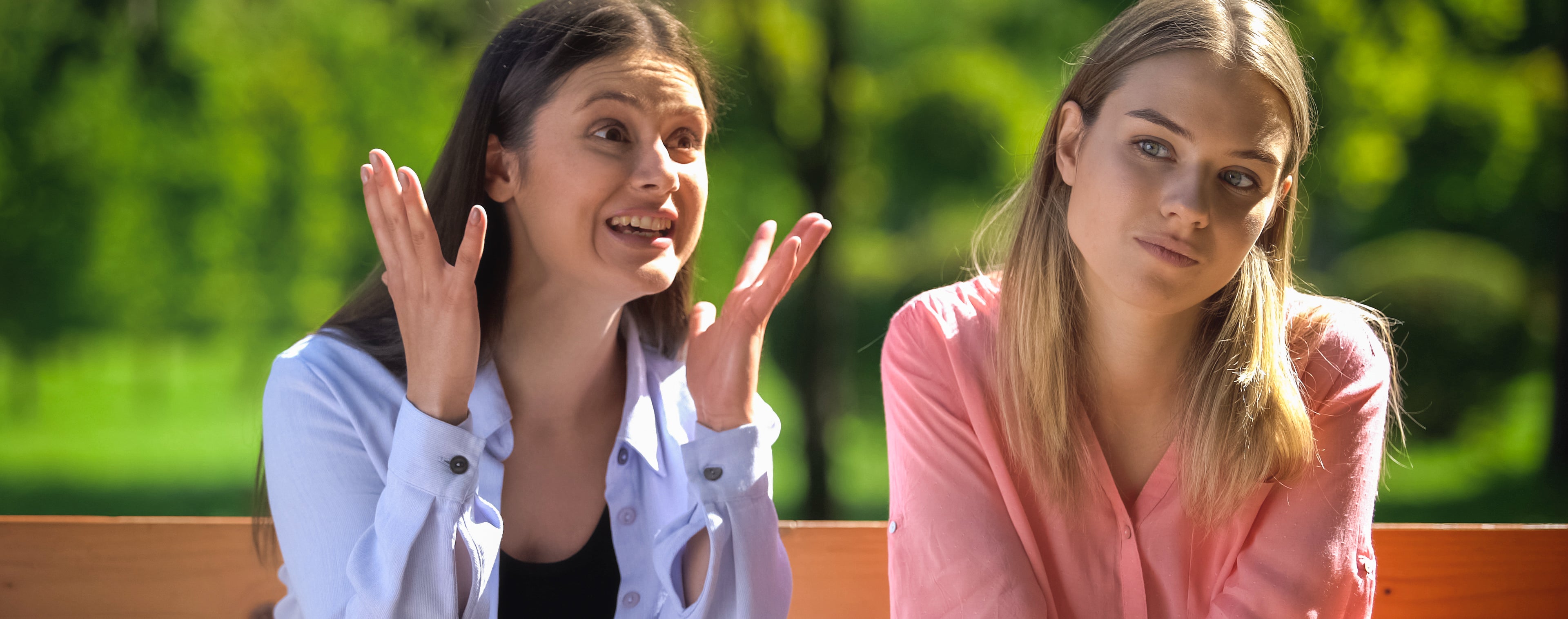 Two women are sitting on a bench outdoors. The woman on the left is talking animatedly, while the woman on the right looks indifferent.