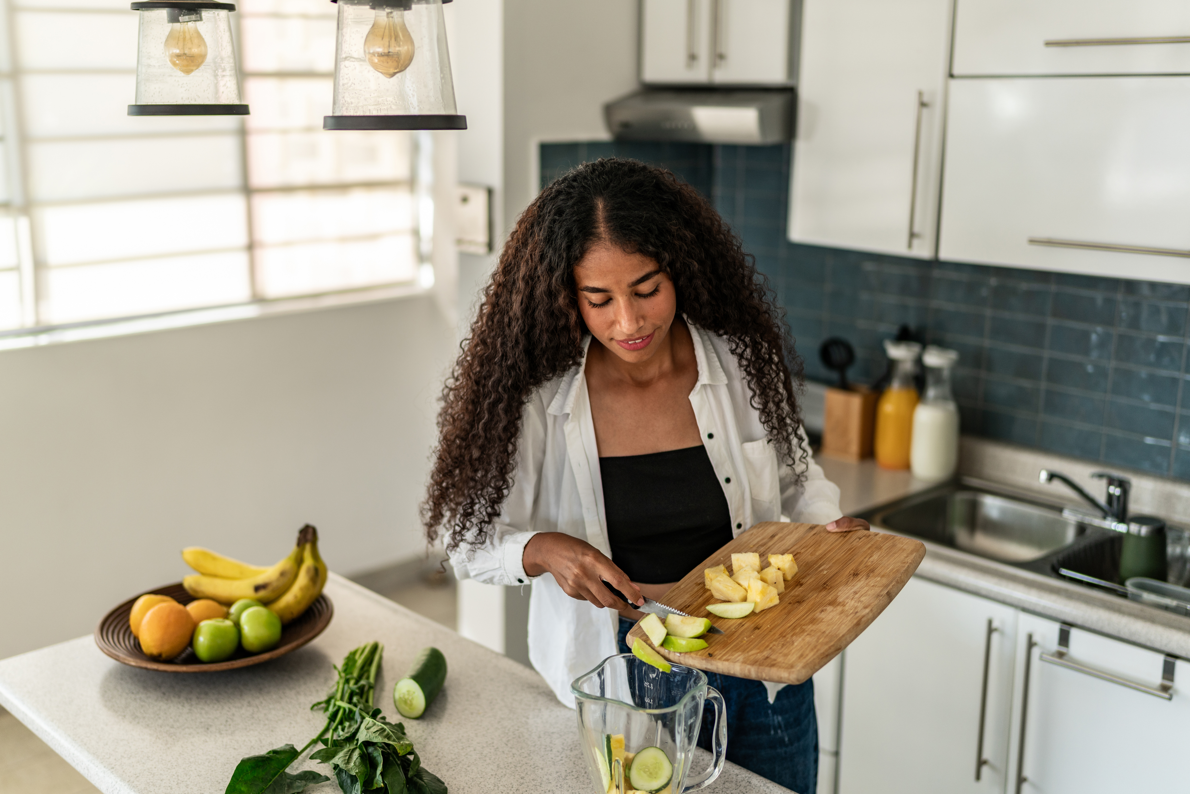 A woman with curly hair in a kitchen slices fruit on a cutting board and adds it to a blender