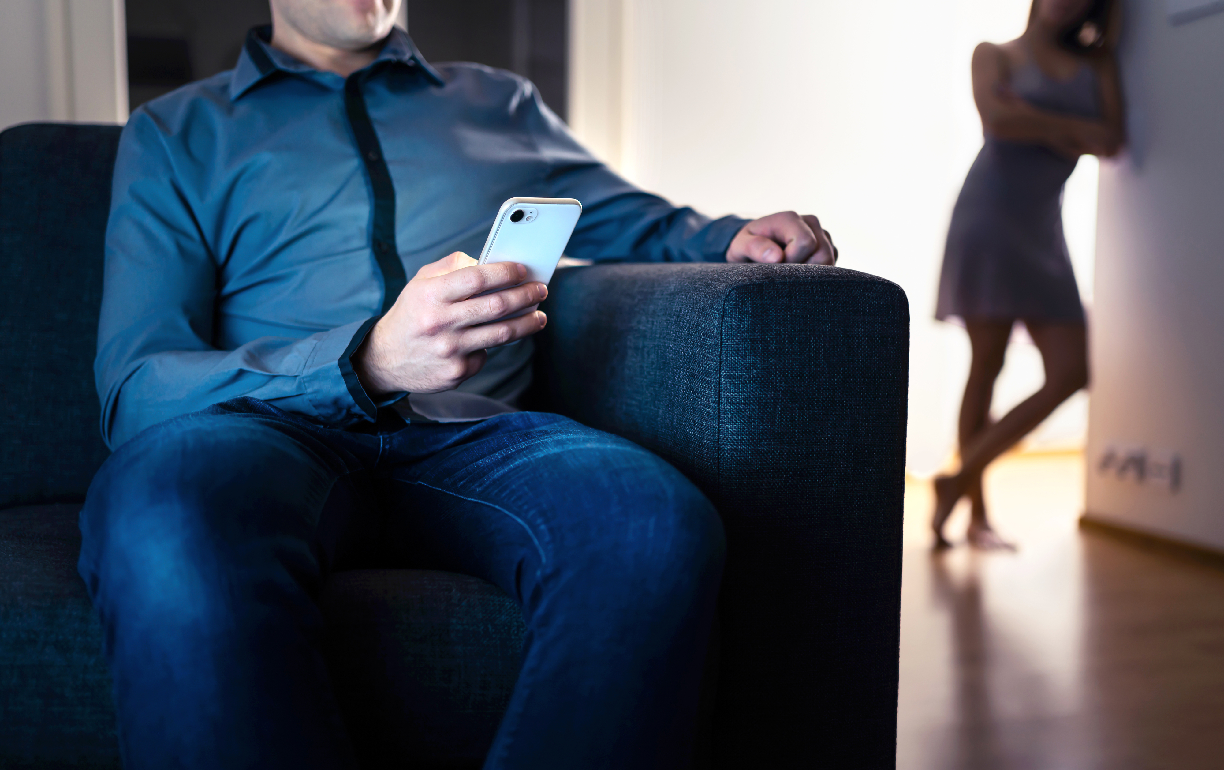 A man sits on a couch looking at his phone while a woman in a dress leans against a wall in the background