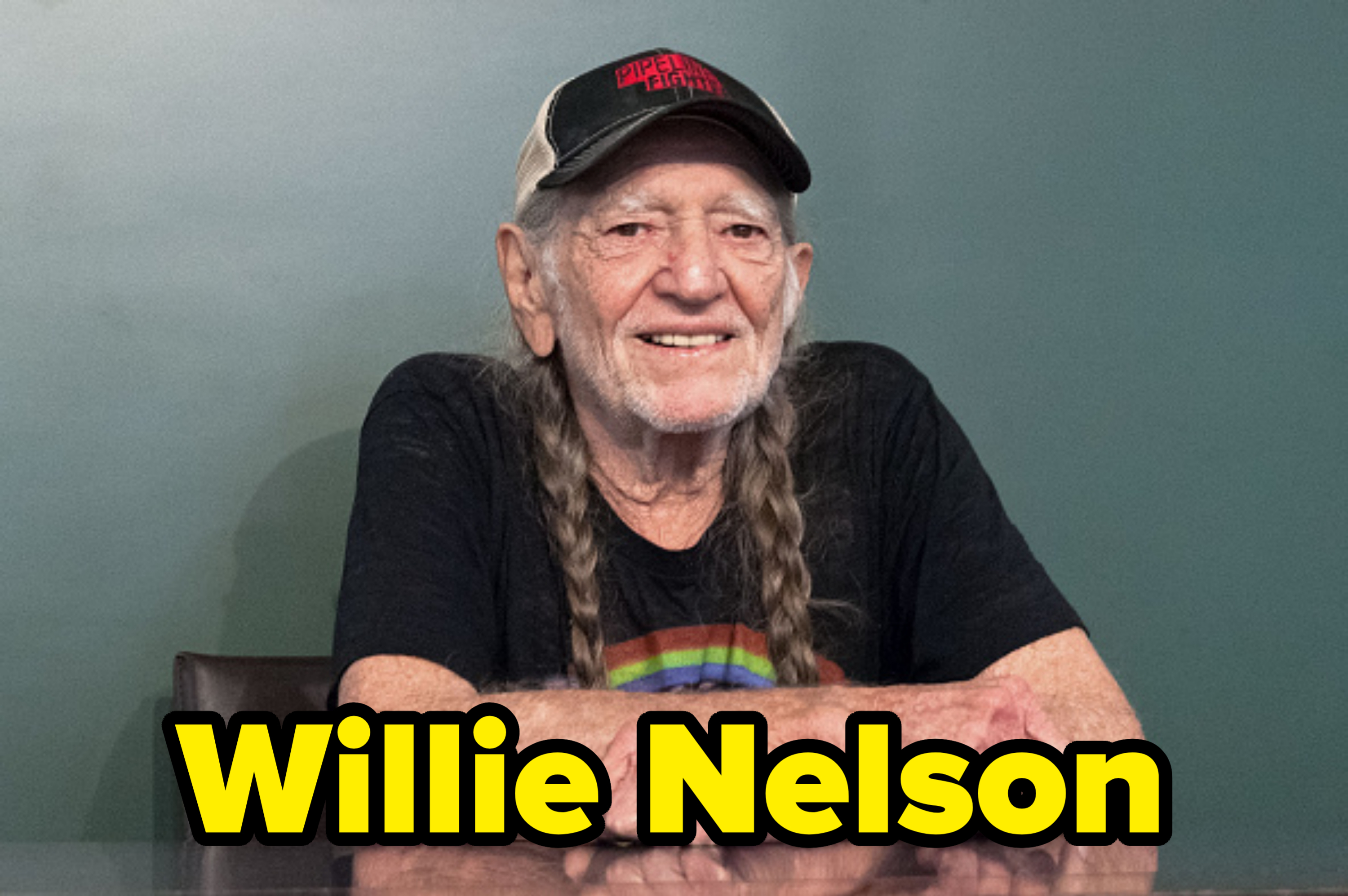 Willie Nelson sits at a table, smiling with his arms crossed. He is wearing a black T-shirt showing a rainbow and a hat with text