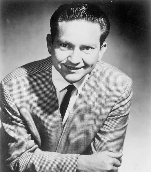 Man in a checkered jacket and tie, smiling and leaning forward with arms crossed