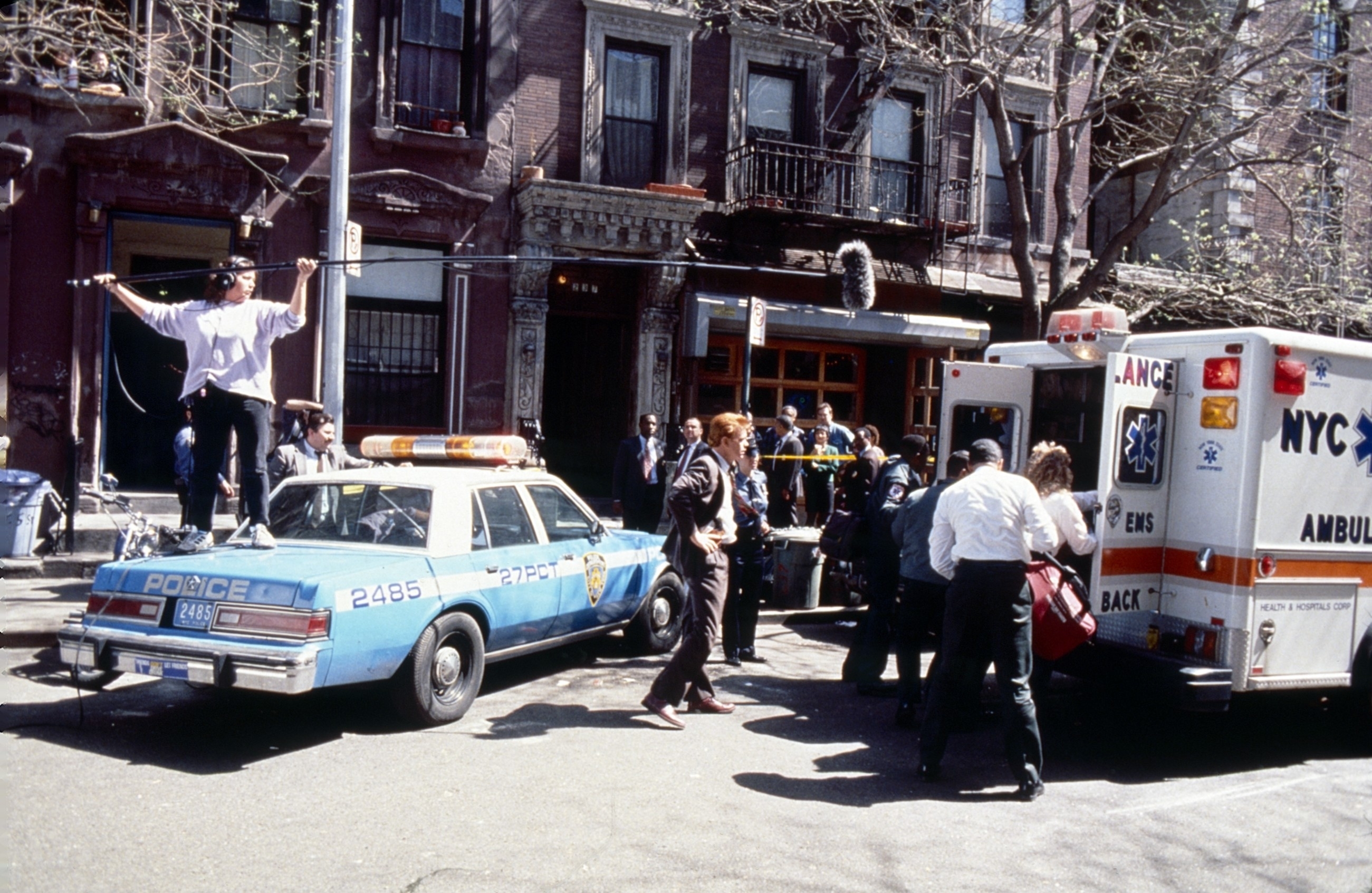 Emergency scene in front of an apartment building in a city, featuring a vintage police car and an ambulance with paramedics attending to people