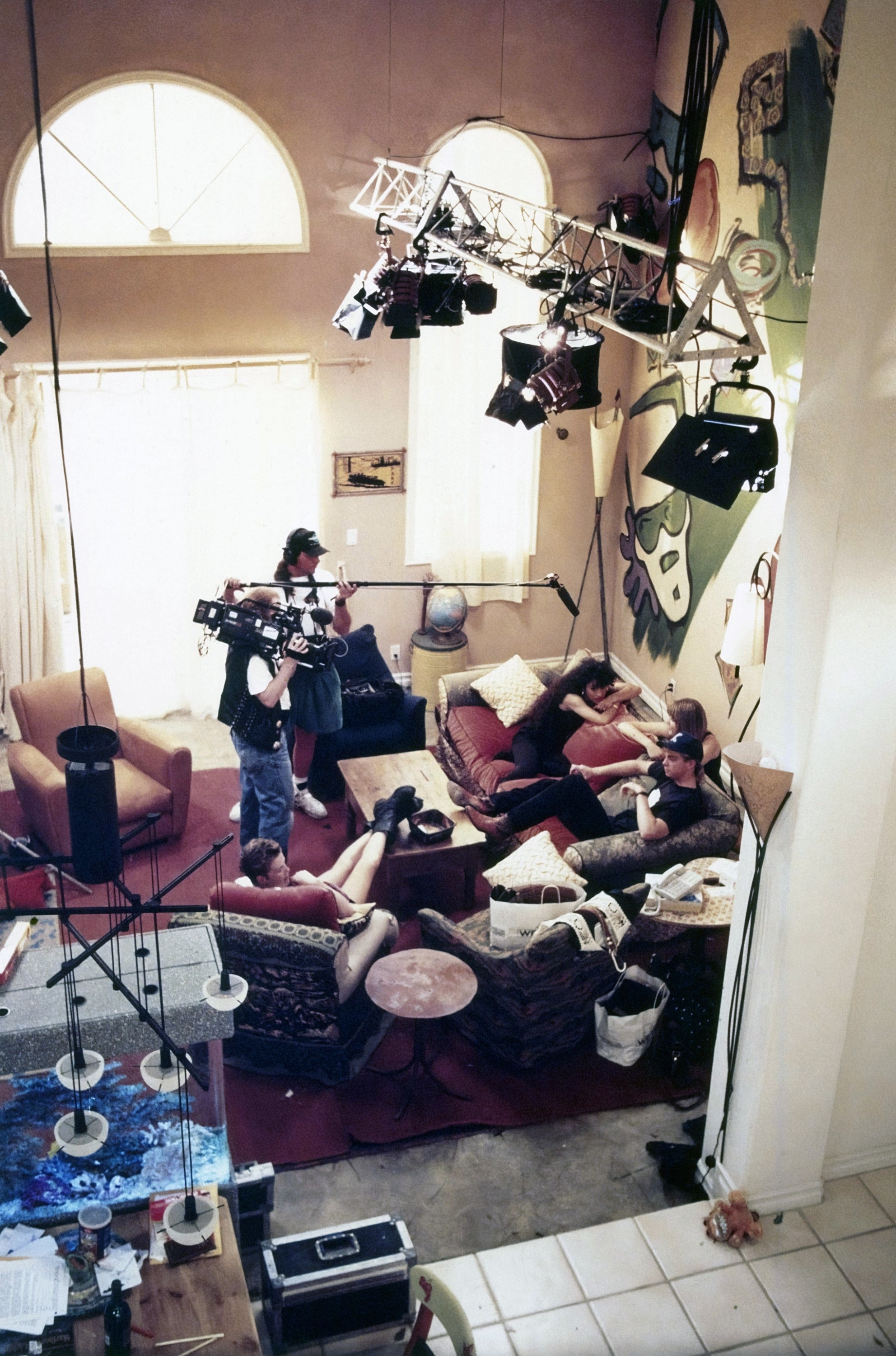 A behind-the-scenes look at a filming set showing a group of crew members working with lights and camera equipment while others relax on the set furniture