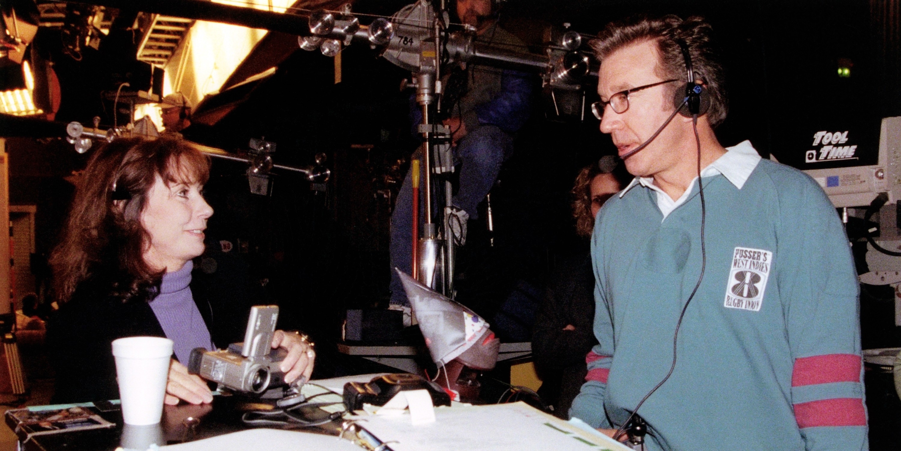 Two people are conversing on a film set. The woman on the left is holding a device, and the man on the right wears a headset and glasses. Various film equipment and props are visible around them