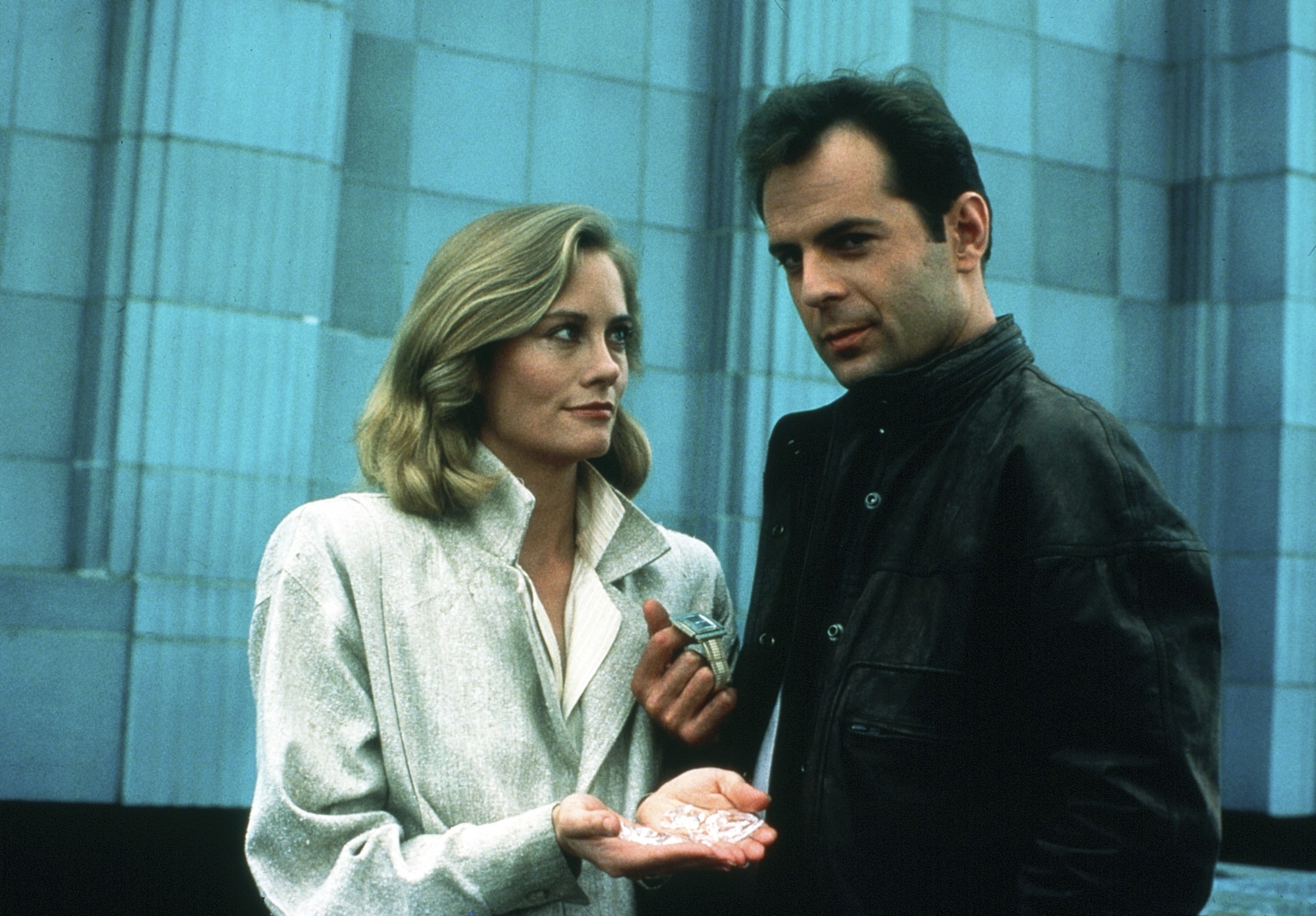 Cybill Shepherd and Bruce Willis stand outside a building. Cybill is holding something in her hand while looking at Bruce, who wears a black jacket