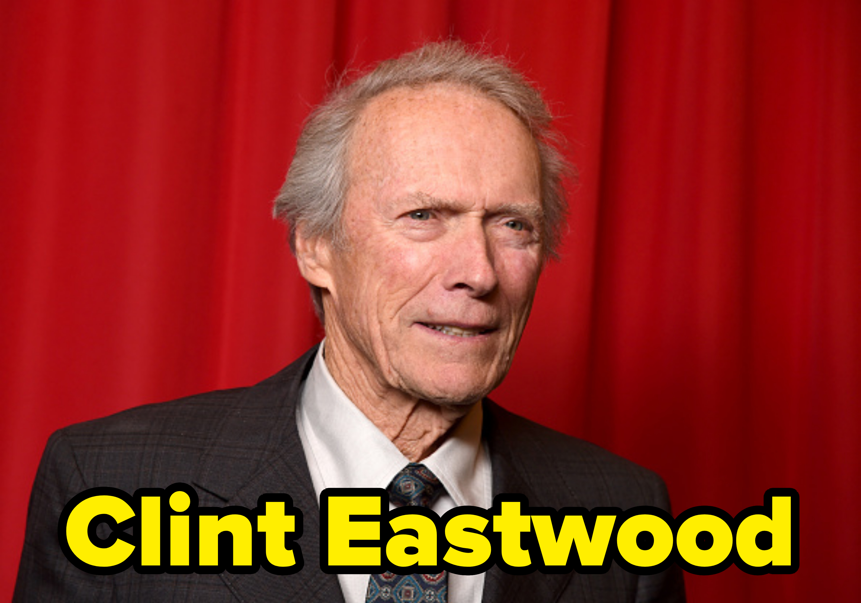 Clint Eastwood in a dark suit and patterned tie, standing in front of a red curtain backdrop