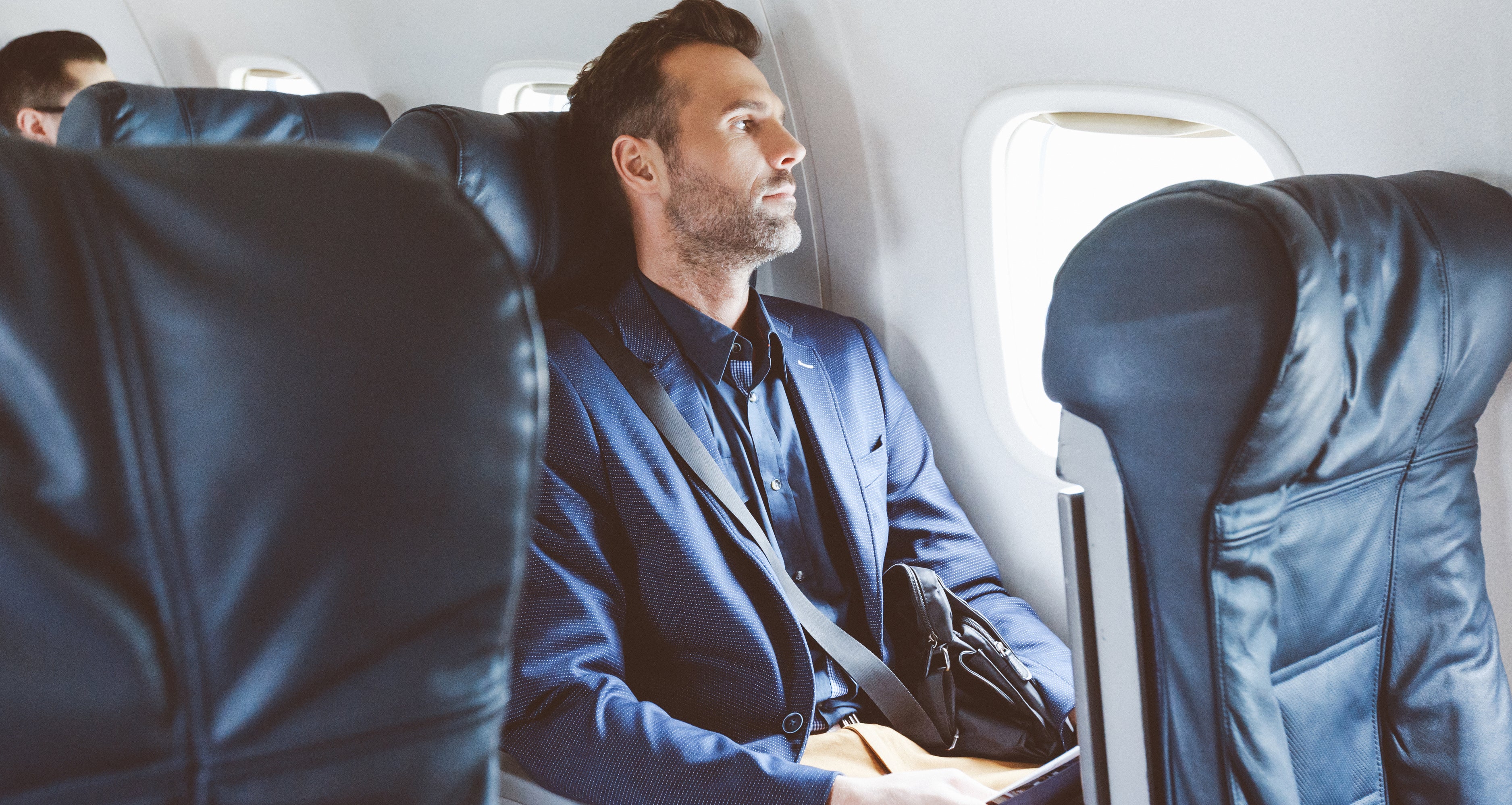 A man sits by the window on an airplane, wearing a blue blazer and holding a tablet.