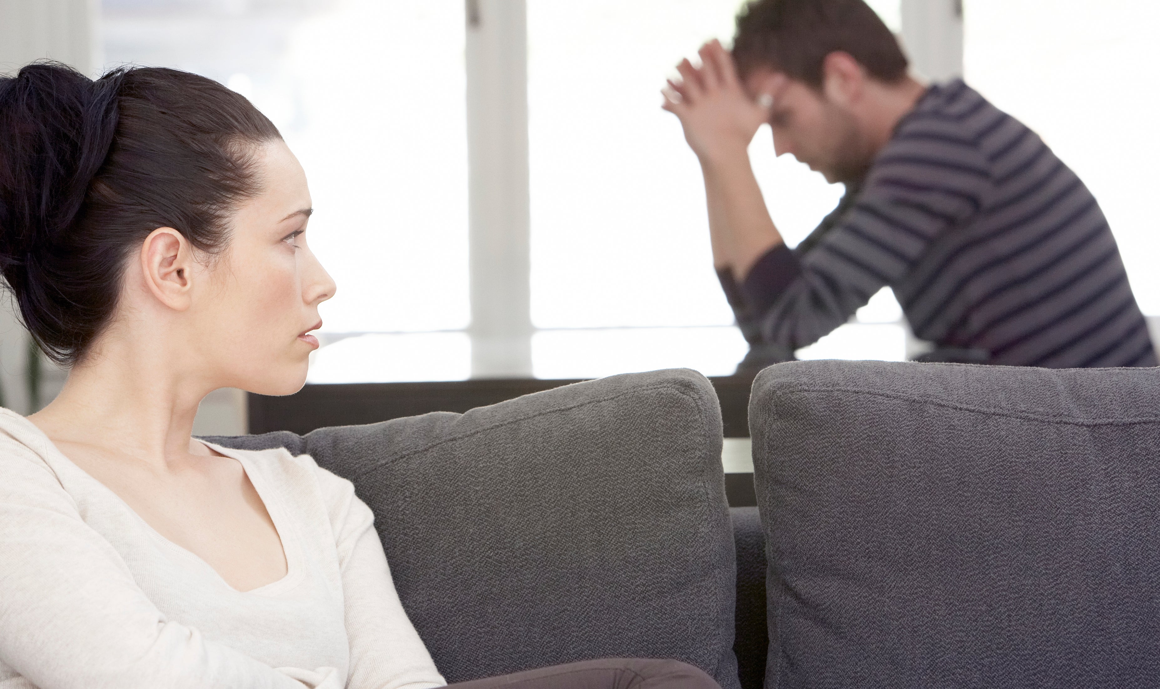 A woman sits on a couch looking to the side with a concerned expression, while a man in the background sits with his head in his hands, appearing distressed
