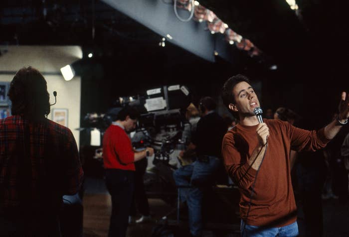 Jerry Seinfeld performing on stage with a microphone, surrounded by a TV crew and camera equipment during the filming of a show