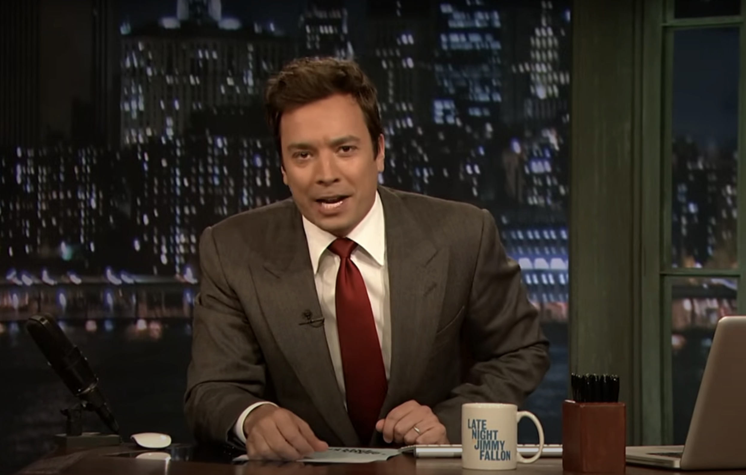 Jimmy Fallon in a suit and tie, seated at a desk on the set of "Late Night with Jimmy Fallon"