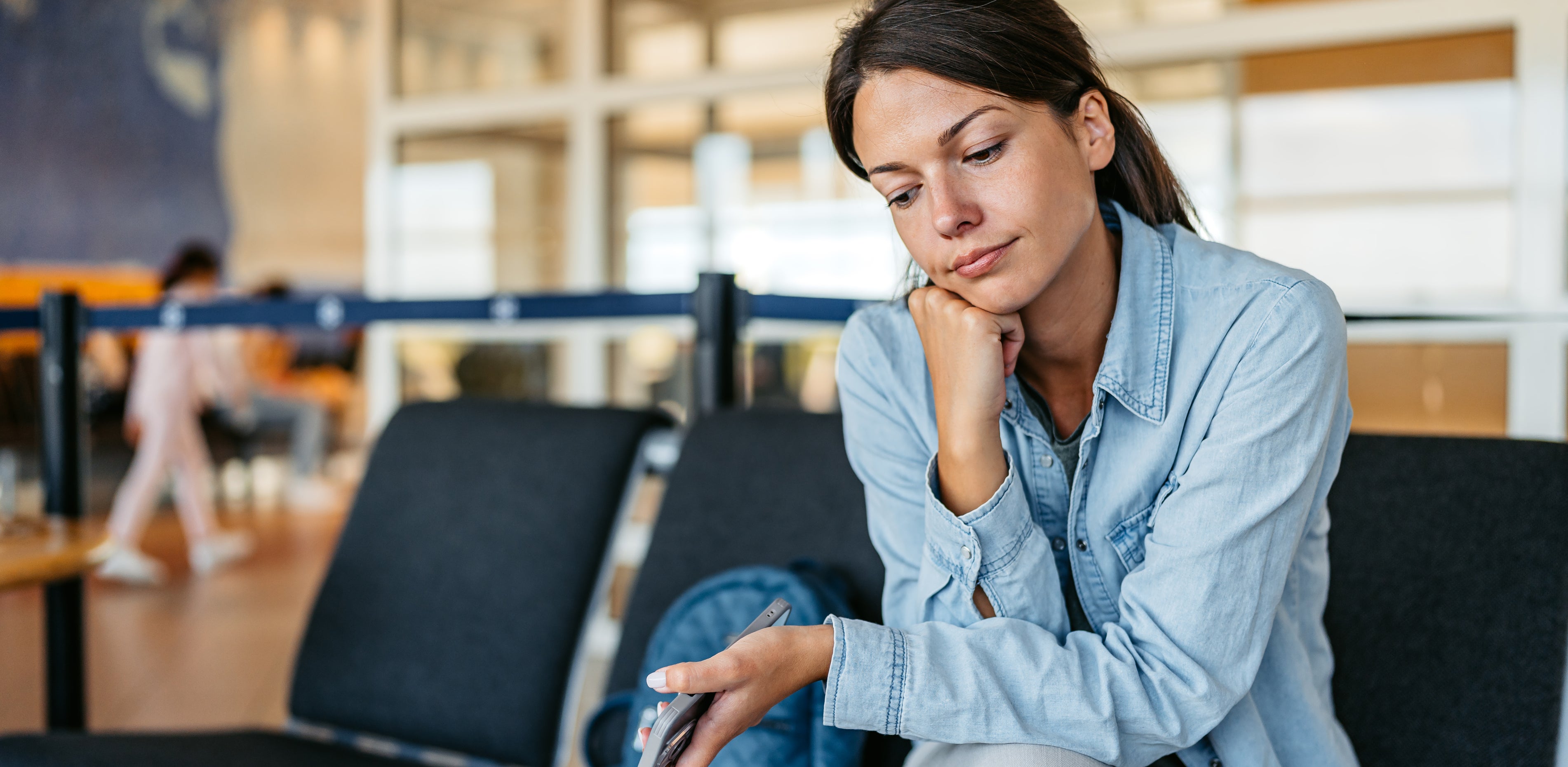 A woman sits in an airport waiting area, resting her head on her hand and looking thoughtful, holding her phone