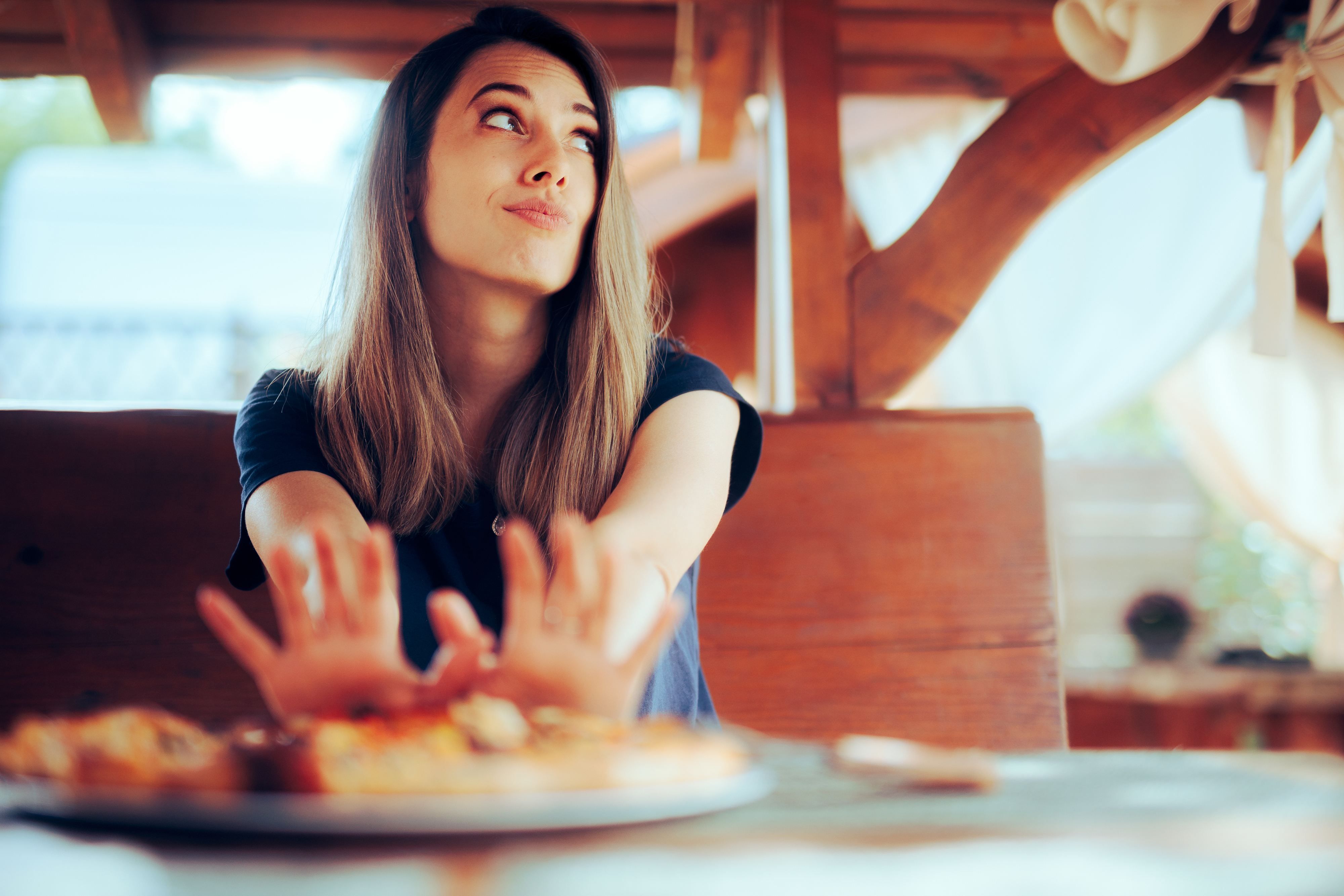 A woman with long hair sits at a wooden table, gesturing with her hands and expressing hesitation or refusal while looking upwards