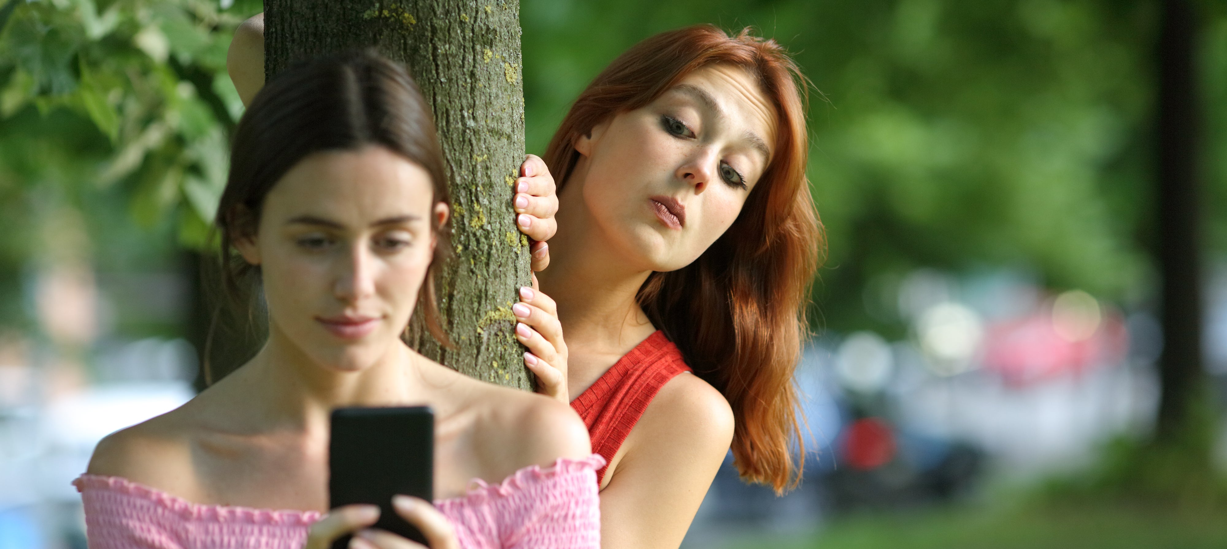 Two women stand by a tree. One is focused on a smartphone, and the other peers curiously from behind the tree
