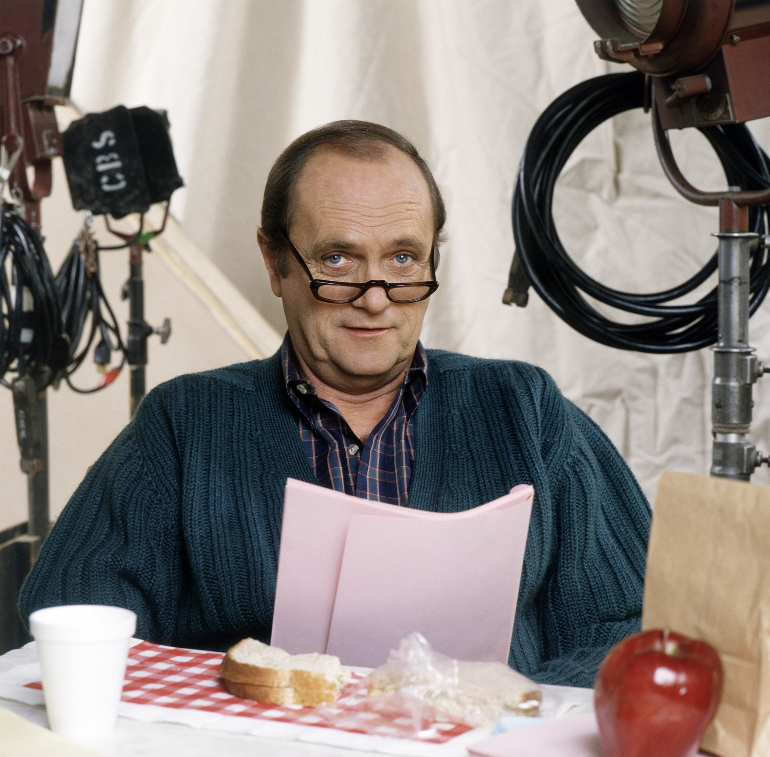 Bob Newhart, wearing glasses and a sweater, reads a script at a table with a sandwich, apple, coffee cup, and film equipment in the background