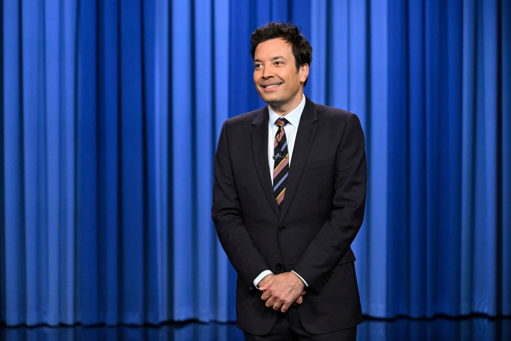Jimmy Fallon stands in front of a blue curtain, wearing a black suit with a striped tie, smiling with hands clasped