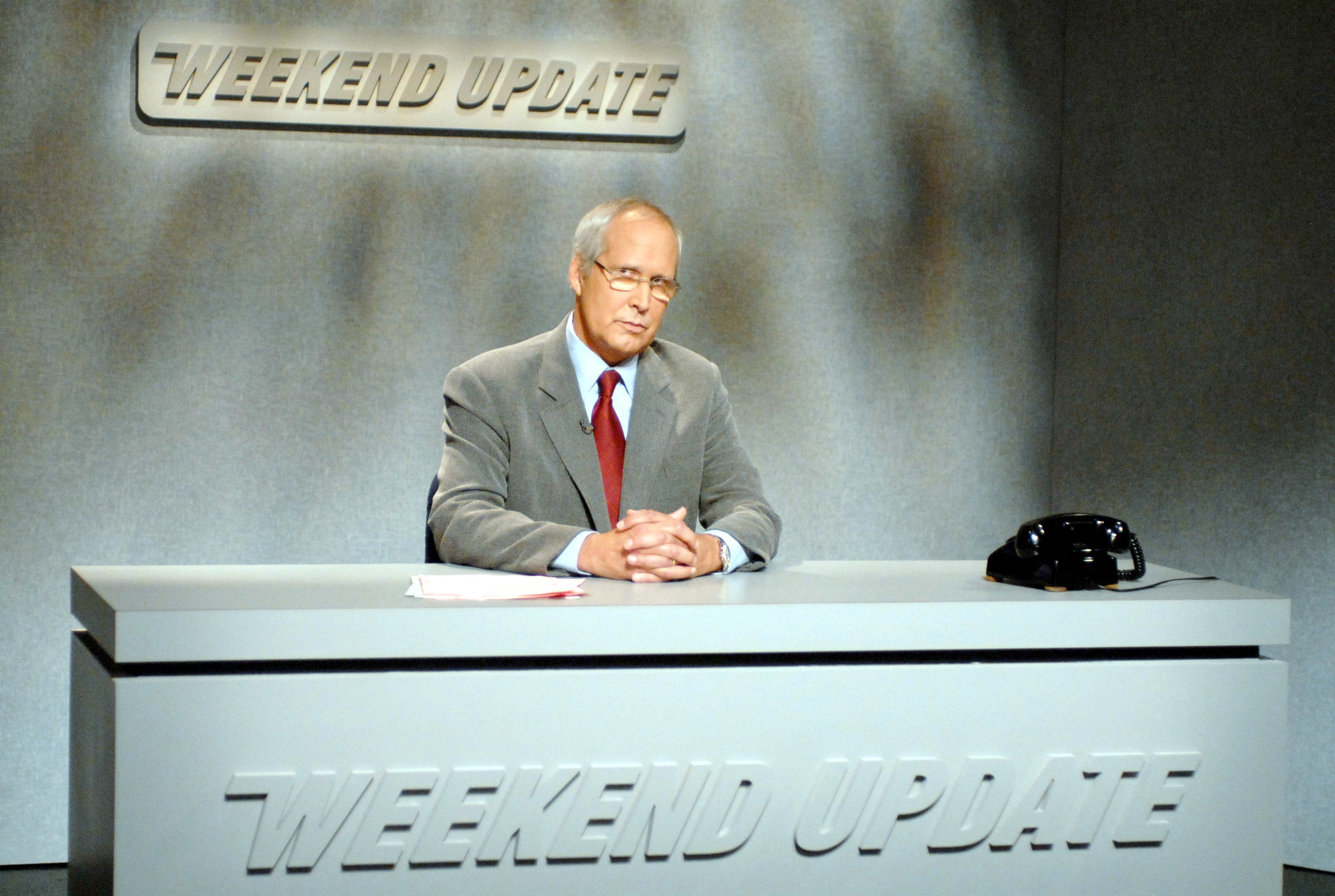 Chevy Chase sits at a "Weekend Update" news desk, wearing a suit and tie, and holding papers while looking at the camera