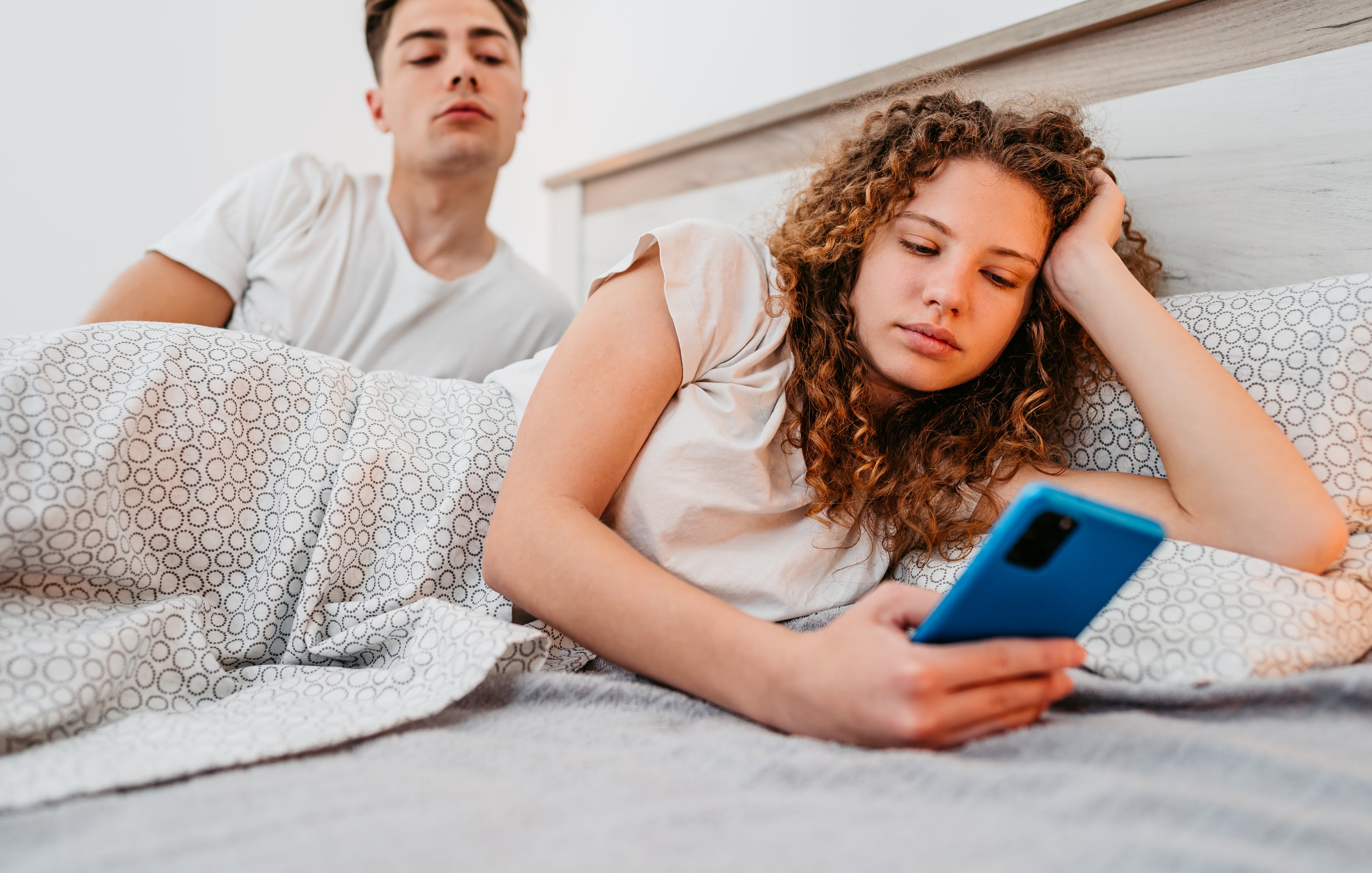 A man and woman lie in bed; the woman looks at her phone while the man, sitting behind her, looks over her shoulder.