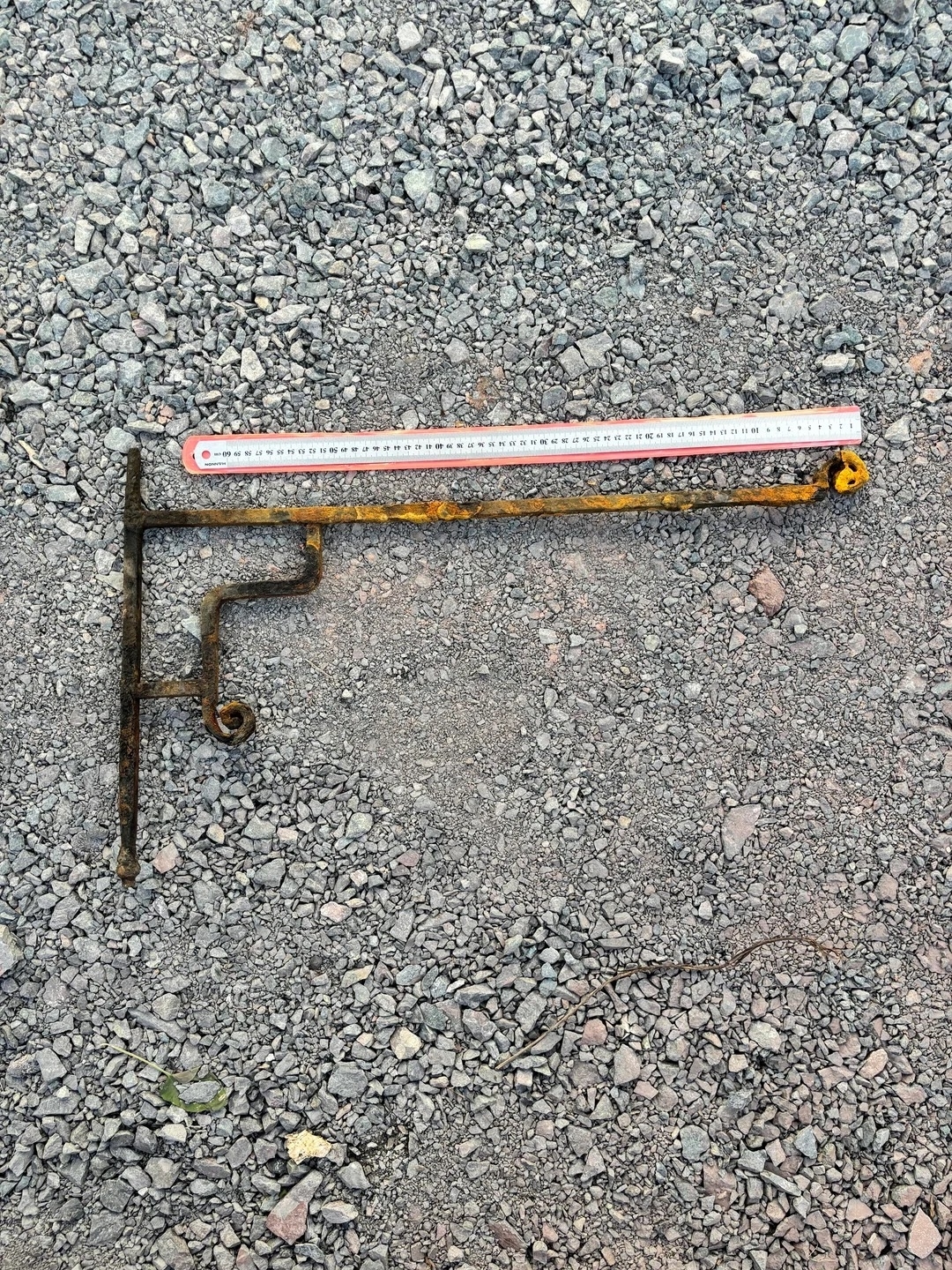 A rusted, L-shaped, metal object with a white measuring tape beside it on a gravel surface