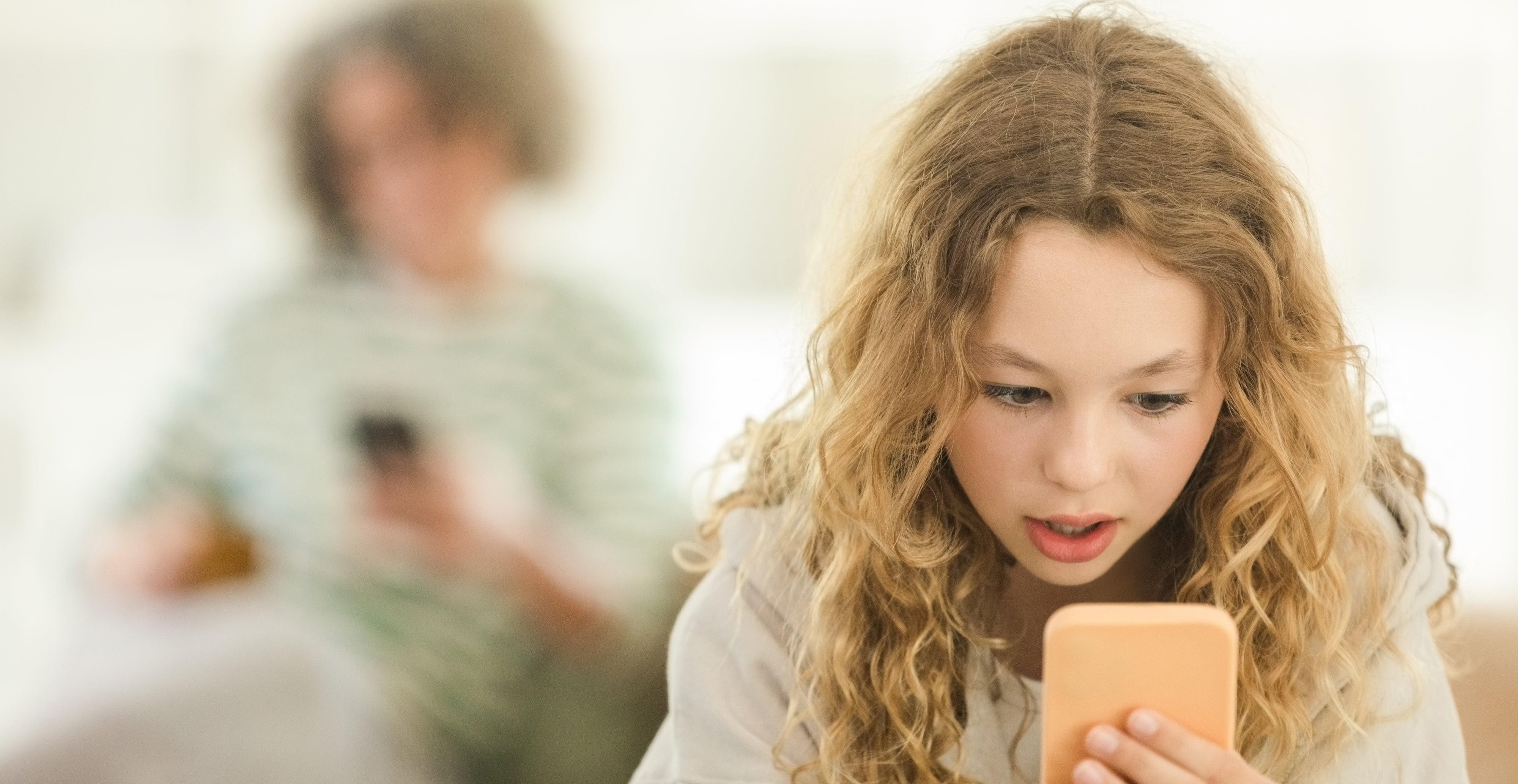 A young girl, with curly hair, looks surprised while holding a smartphone. An older person in the background also uses a phone